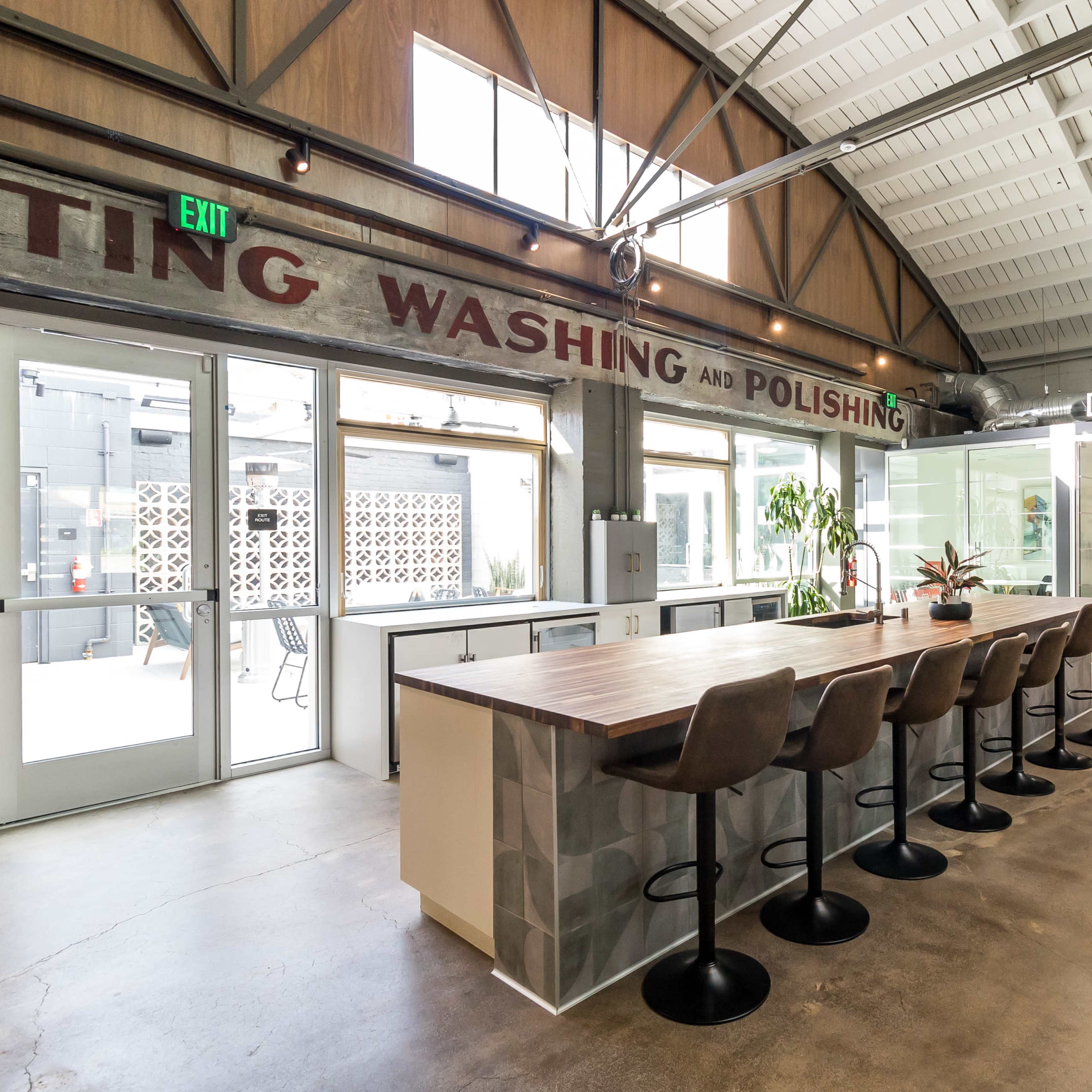 An industrial-style workspace with a long wooden table and bar stools, along with large windows and modern amenities.