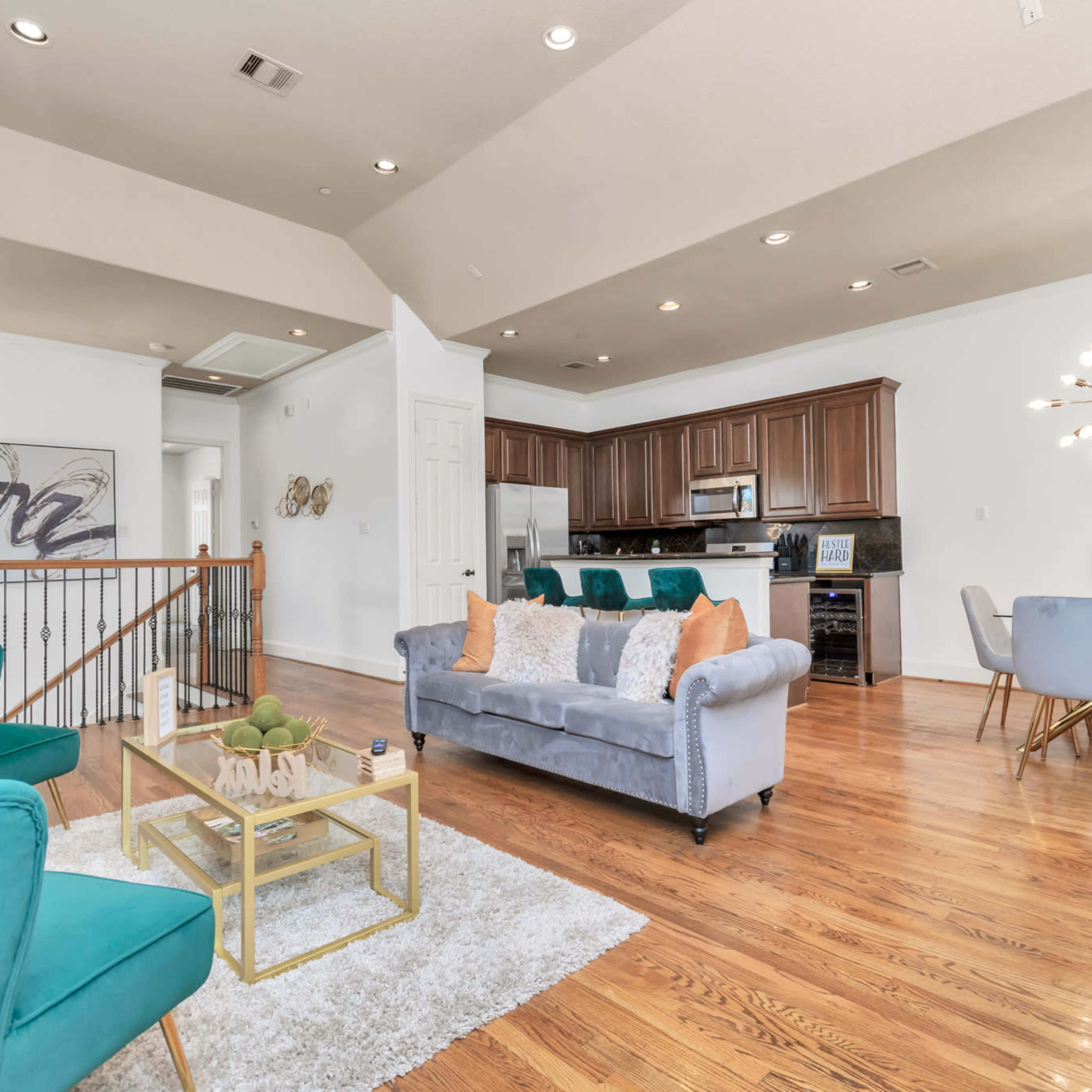 A modern open-concept living area featuring a blue couch, teal accent chairs, and a dining table, with hardwood floors and a kitchen in the background.