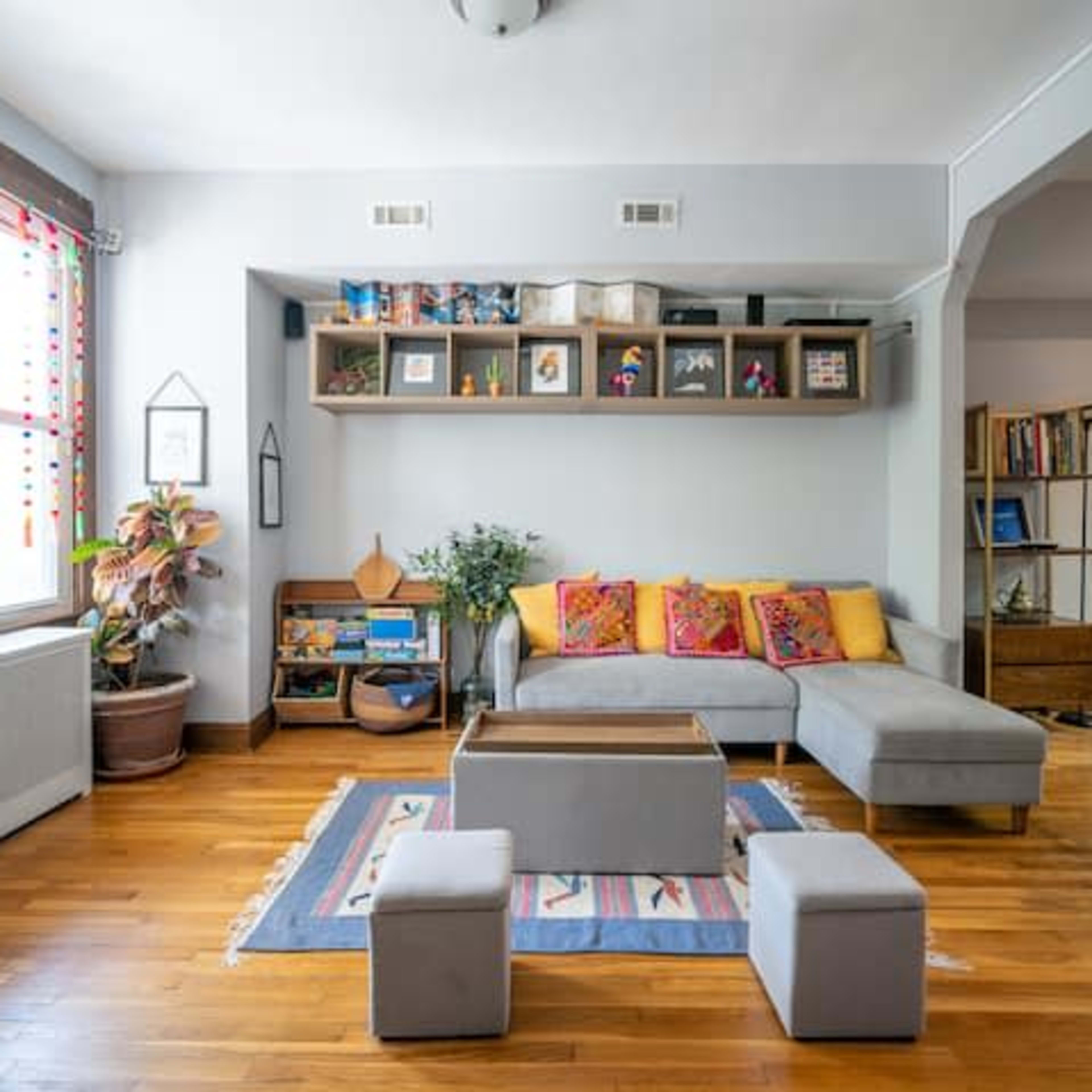 The living room features a gray sectional sofa with colorful pillows, a wooden coffee table, and a bookshelf against the wall, with two small stools on a patterned rug.