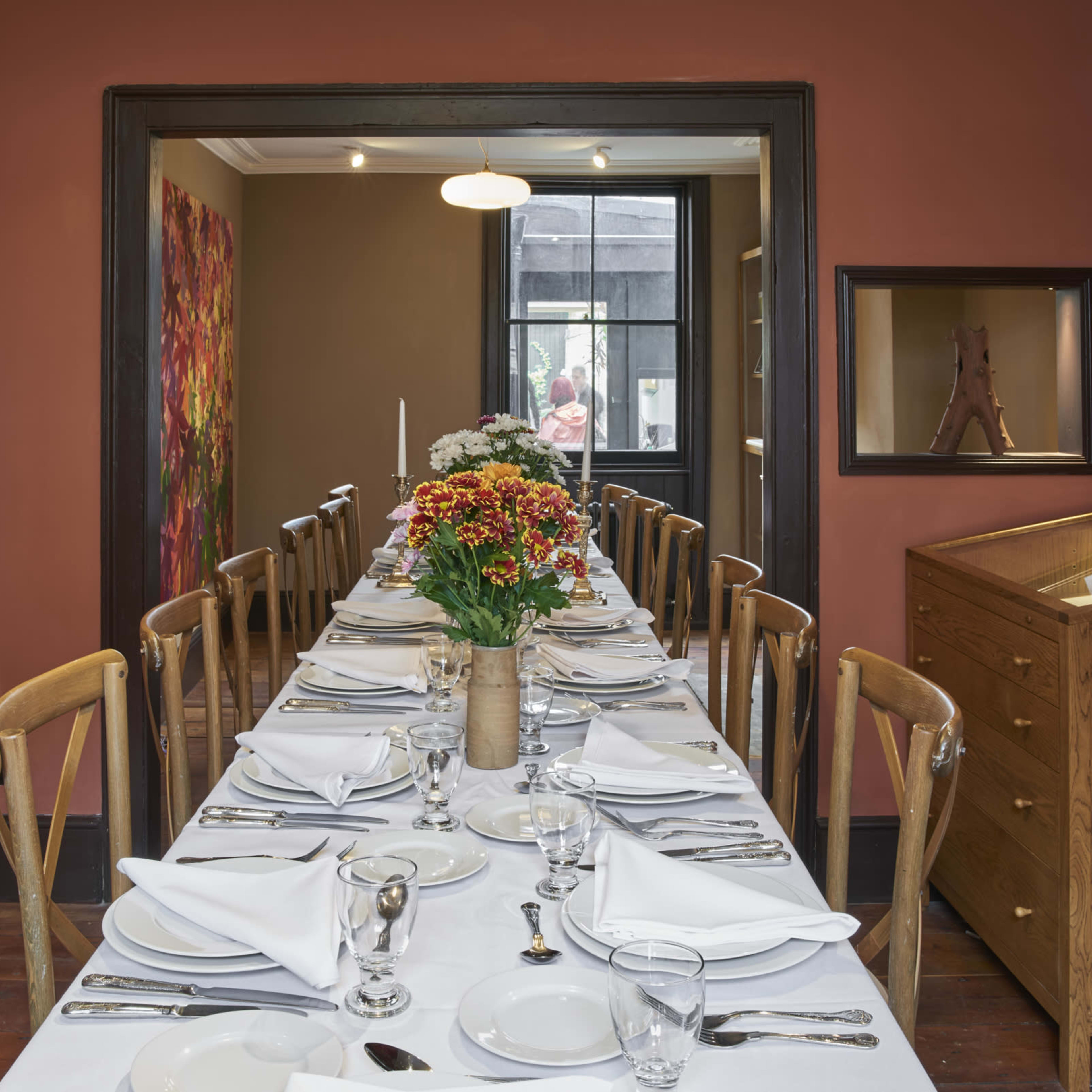 A long dining table is set with white linens, utensils, and a floral centerpiece in a warmly colored room with decorative art on the walls.