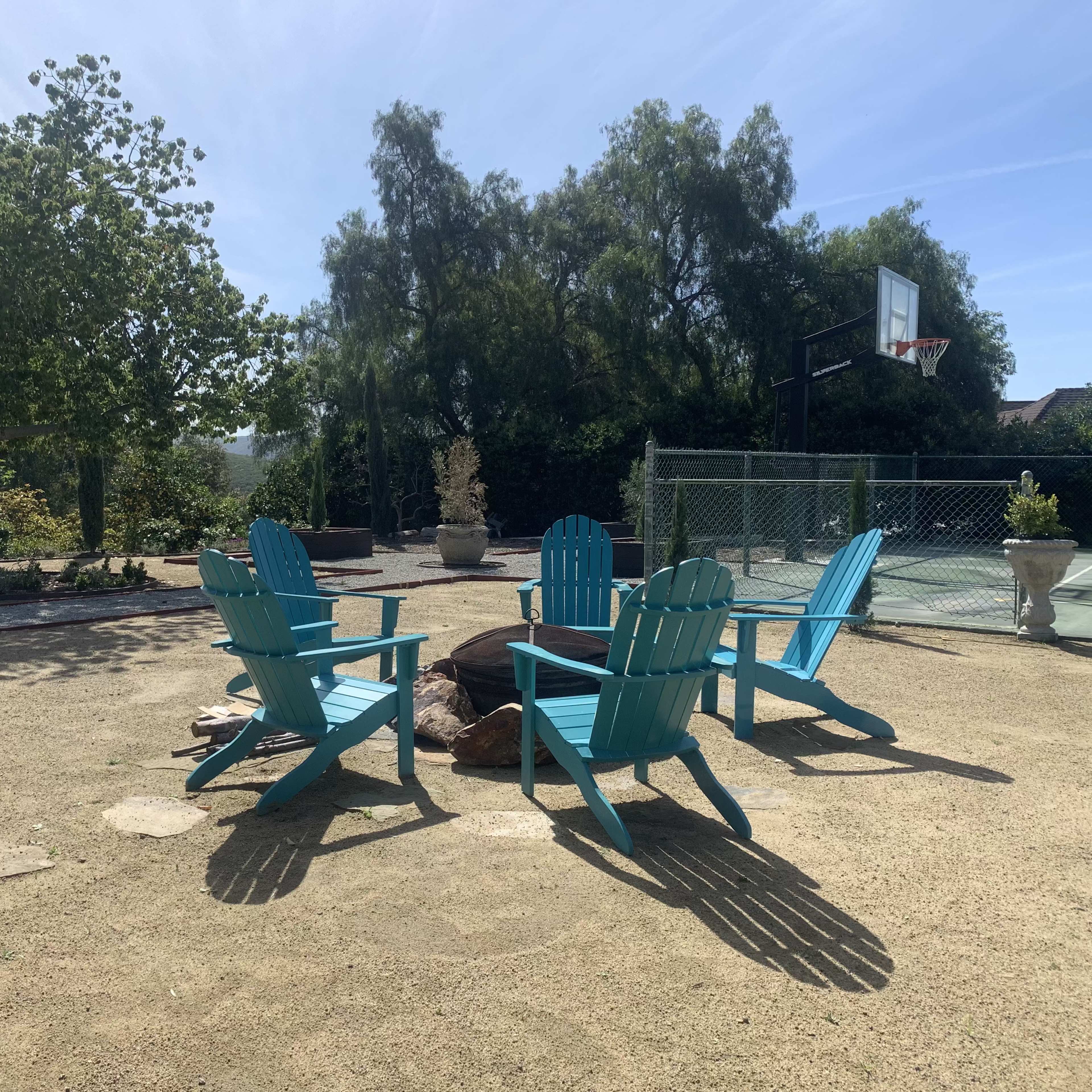 A circular arrangement of blue Adirondack chairs around a small fire pit in a landscaped outdoor area with a basketball hoop in the background.