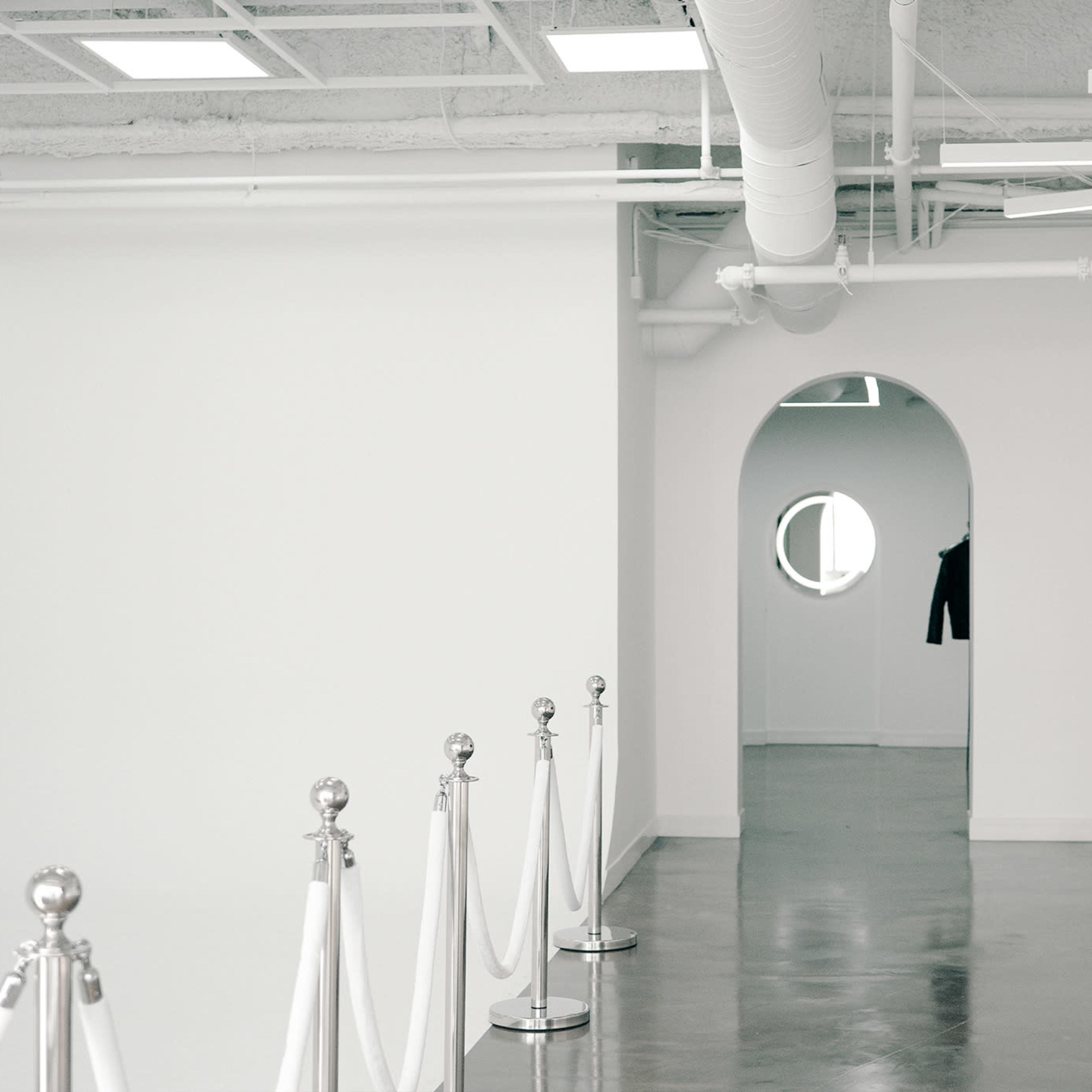 A modern, minimalist interior with white walls, a rounded entrance, a circular window, and a long, rectangular white counter, lined by stanchions and ropes.