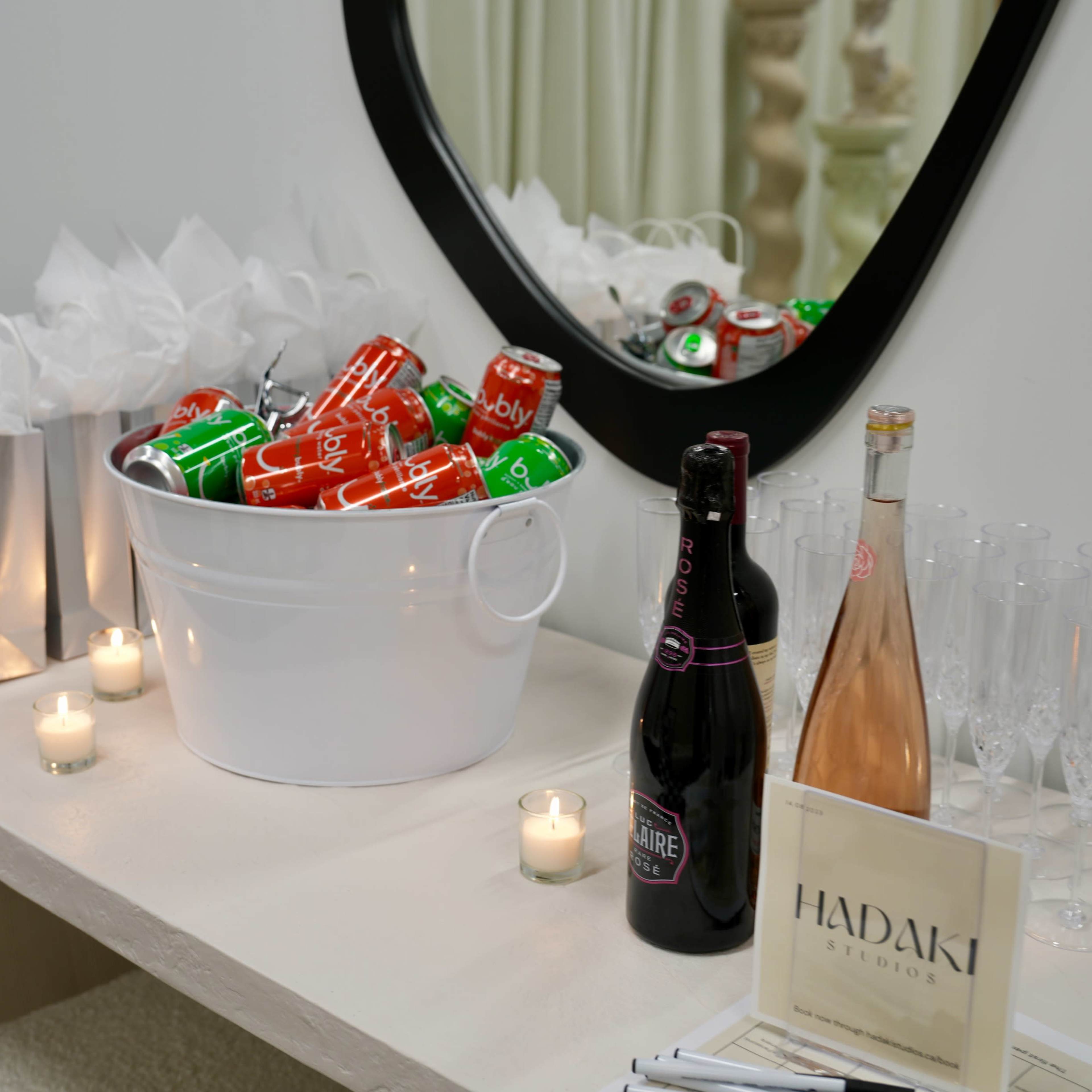 A white bucket filled with soft drink cans sits on a table beside bottles of sparkling wine and candles, with gift bags and a sign displayed nearby.