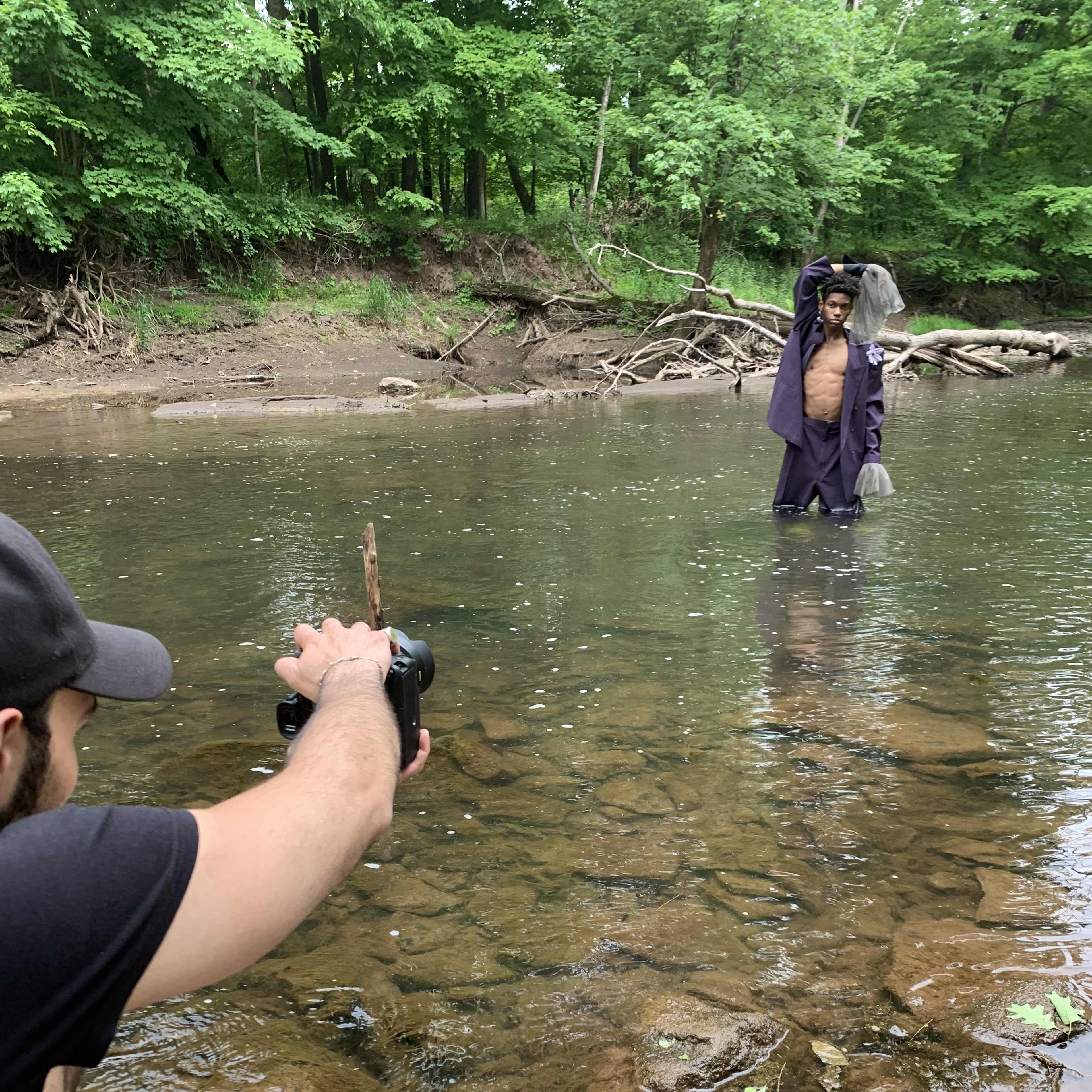 A person stands in a shallow stream while another person takes a photograph from the shore.