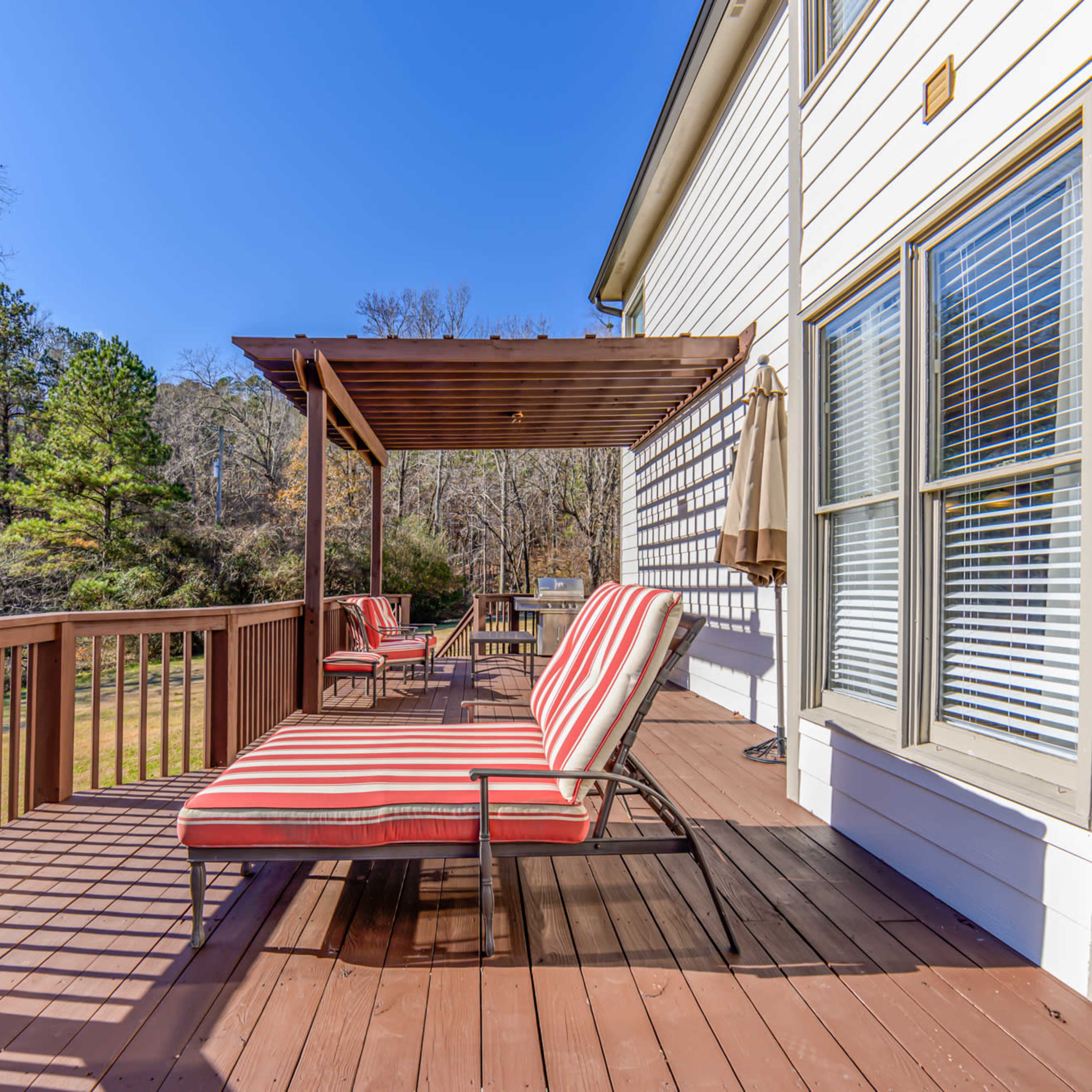 A wooden deck features two striped lounge chairs under a pergola beside a house, with trees visible in the background.