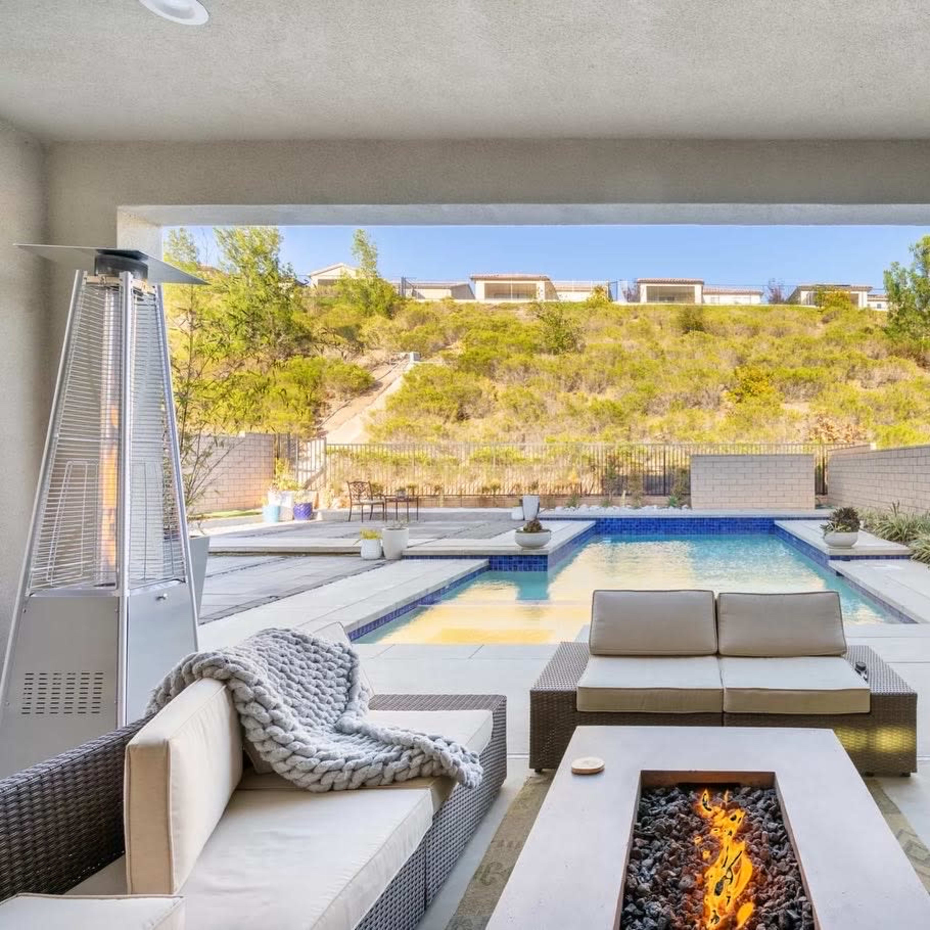 A modern outdoor lounge area features a fire pit and seating beside a pool with a hillside backdrop.