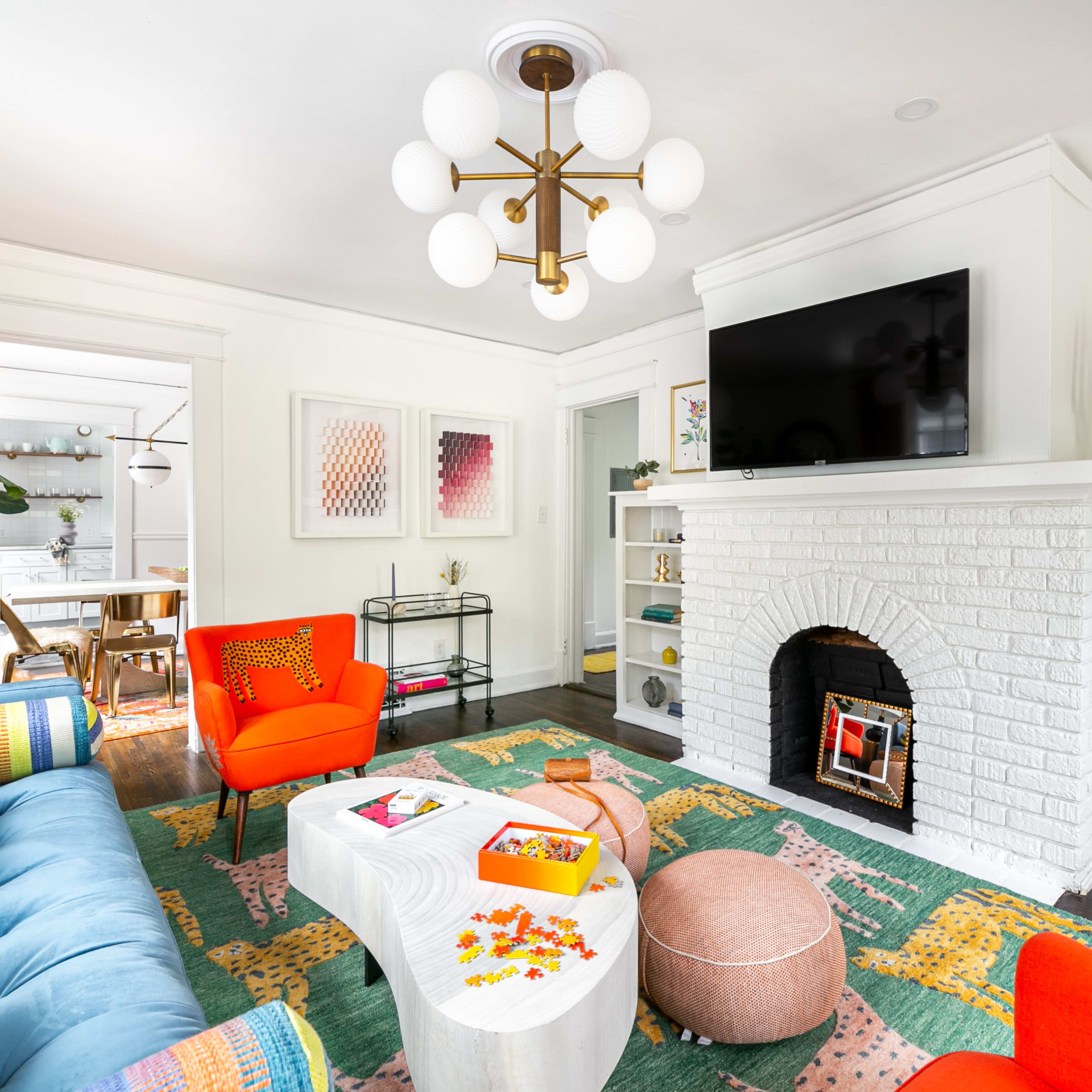 A living room features a colorful sofa, bright chairs, a white brick fireplace with a mounted television, and a coffee table surrounded by decorative items and plants.