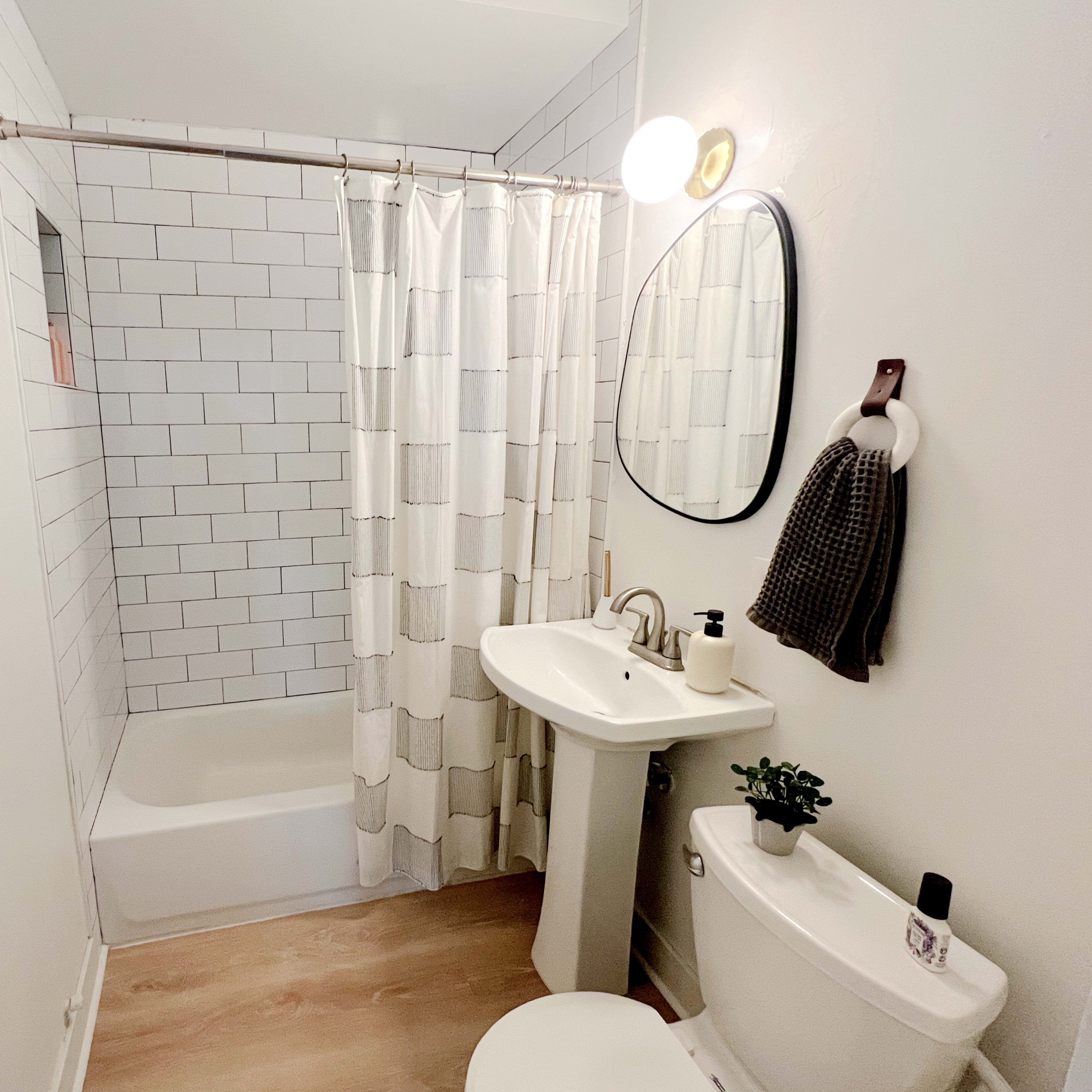 The bathroom features a bathtub with a shower curtain, a pedestal sink, a round mirror, and minimalist decor in a clean layout.