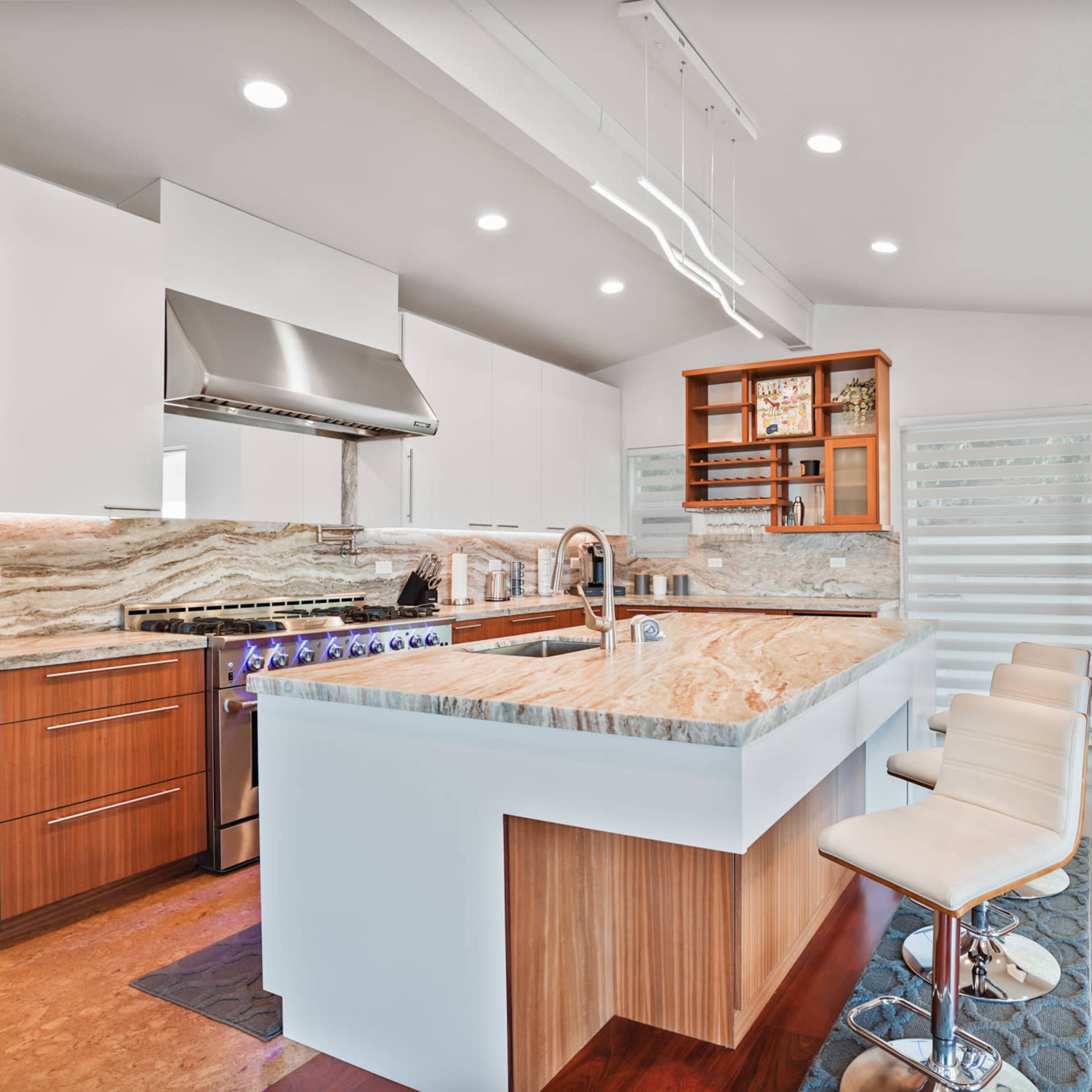A modern kitchen features sleek cabinetry, a large stone countertop, and bar seating, illuminated by recessed lights.