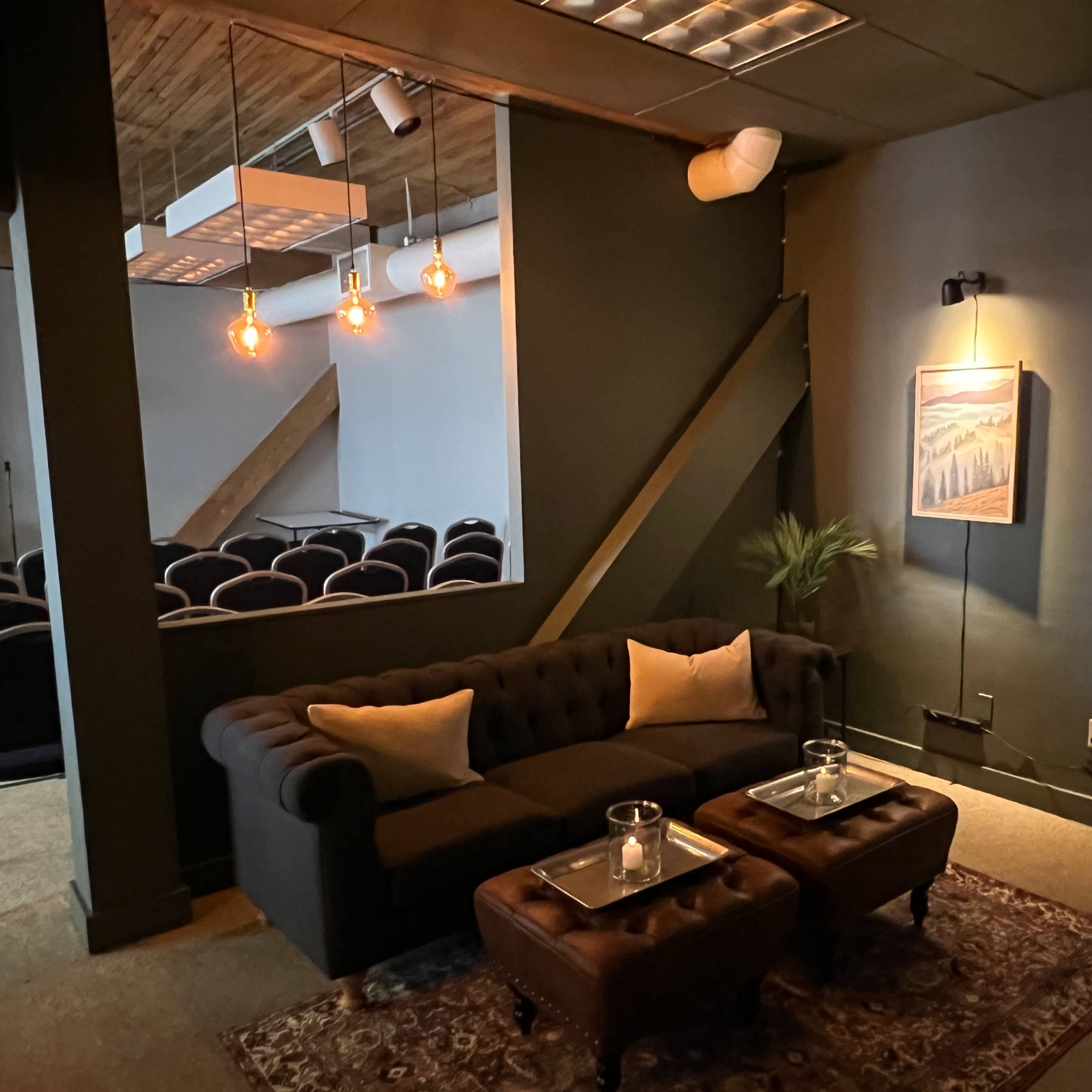 A cozy lounge area features a dark sofa with cushions, two wooden tables, and soft lighting, leading to a seating area in the background.