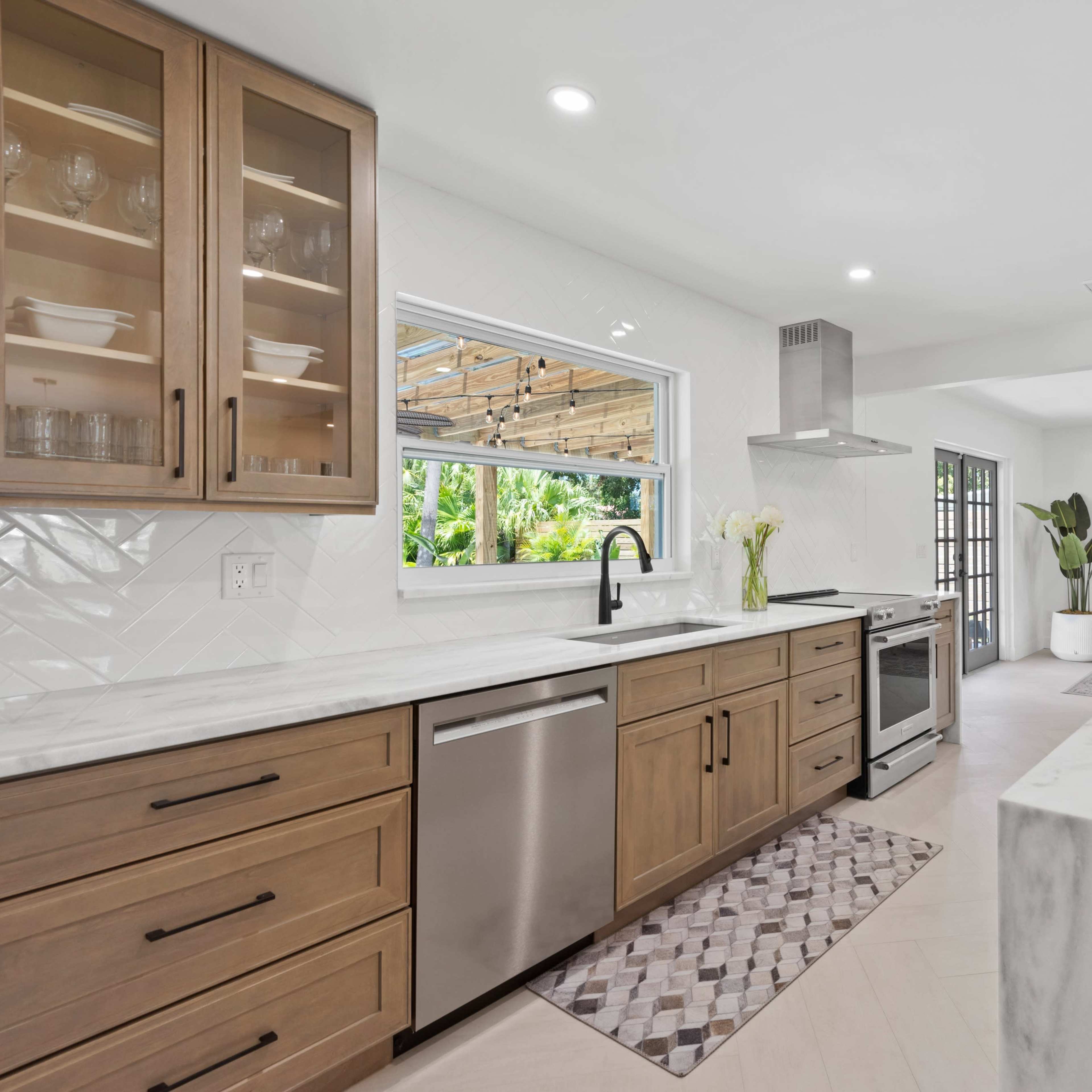 A modern kitchen featuring wooden cabinets, stainless steel appliances, a marble countertop, and a view to an outdoor area with greenery.