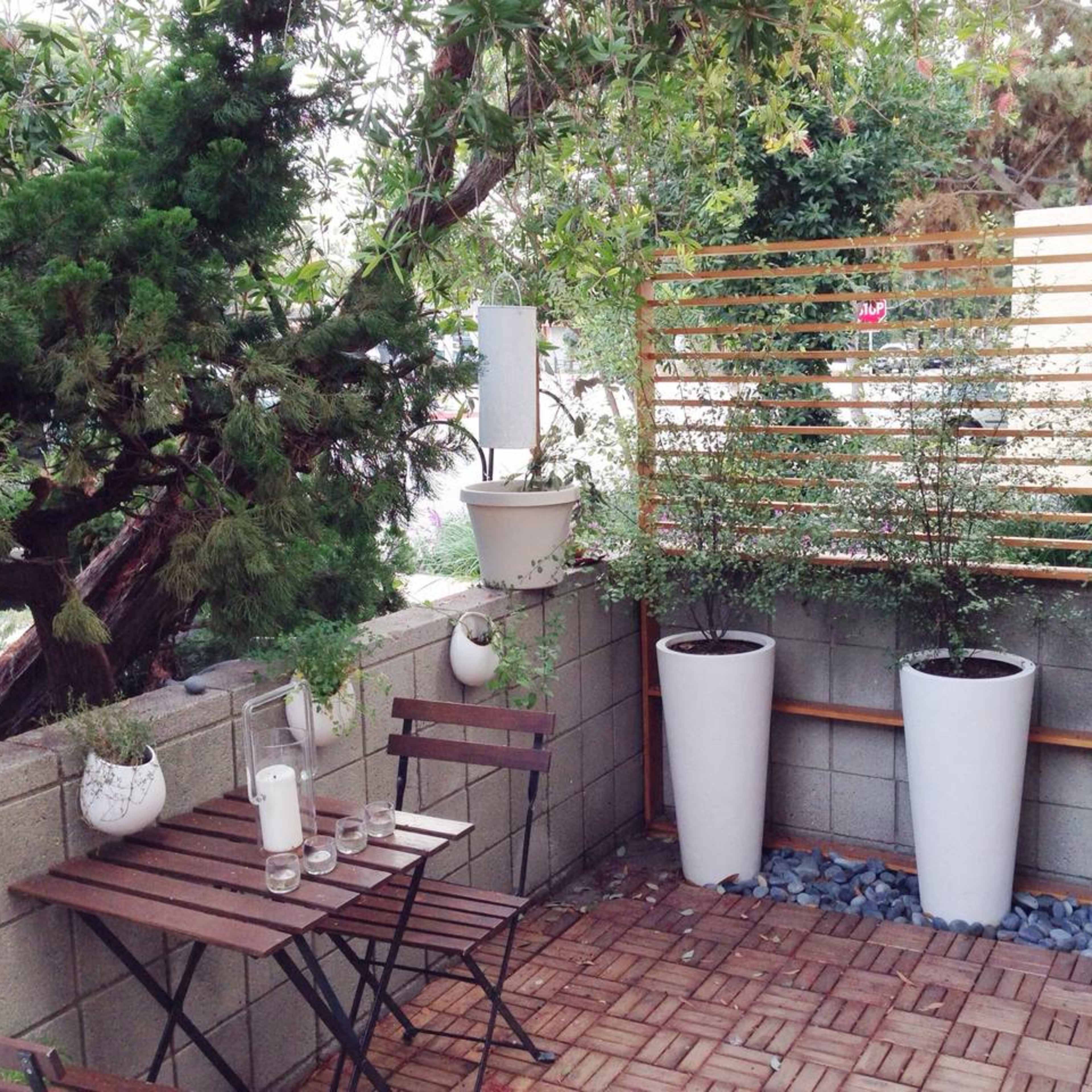 A small outdoor garden area featuring a wooden table and chairs, along with potted plants against a textured wall.