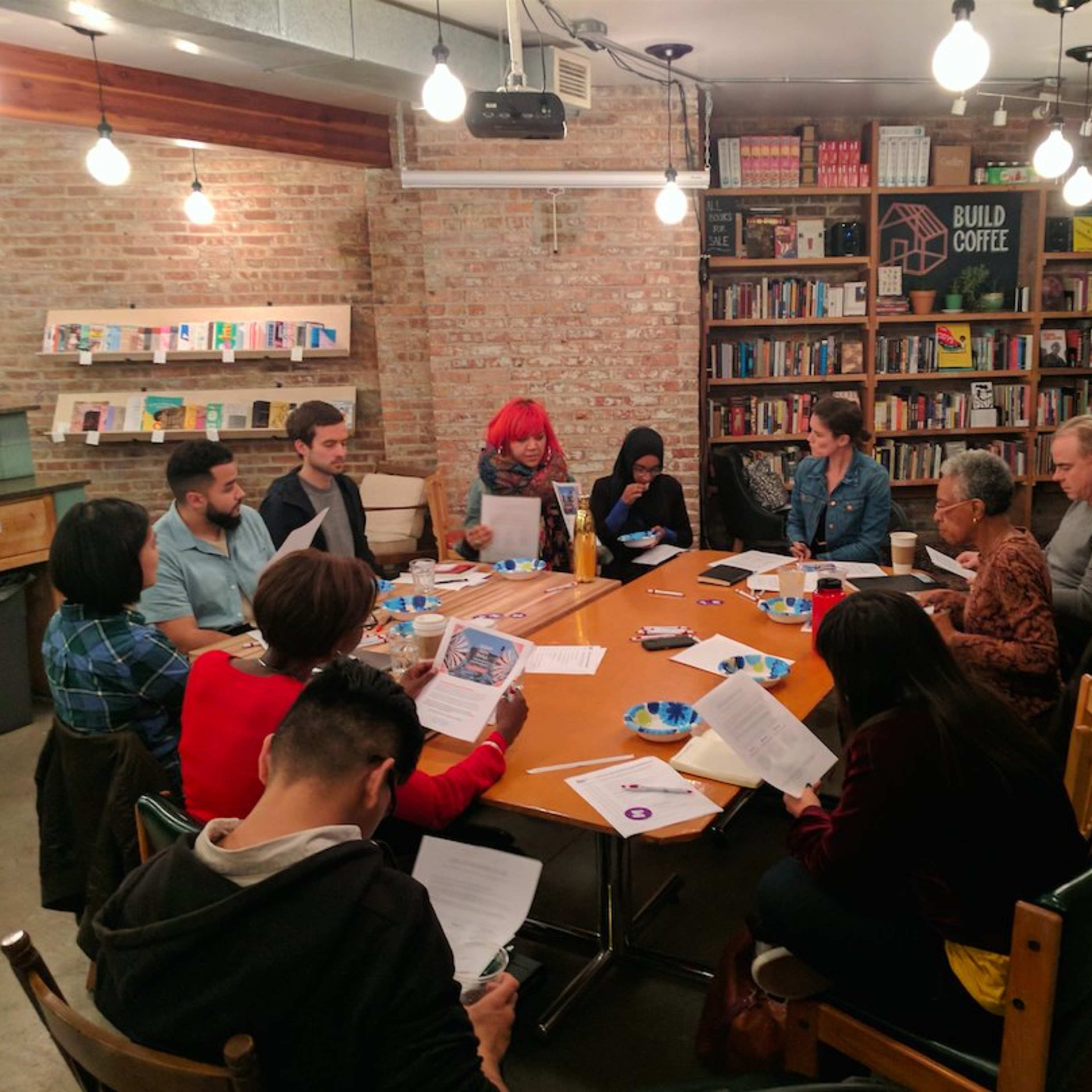 A group of diverse individuals is seated around a large table in a cozy, book-lined space, participating in a meeting or discussion.