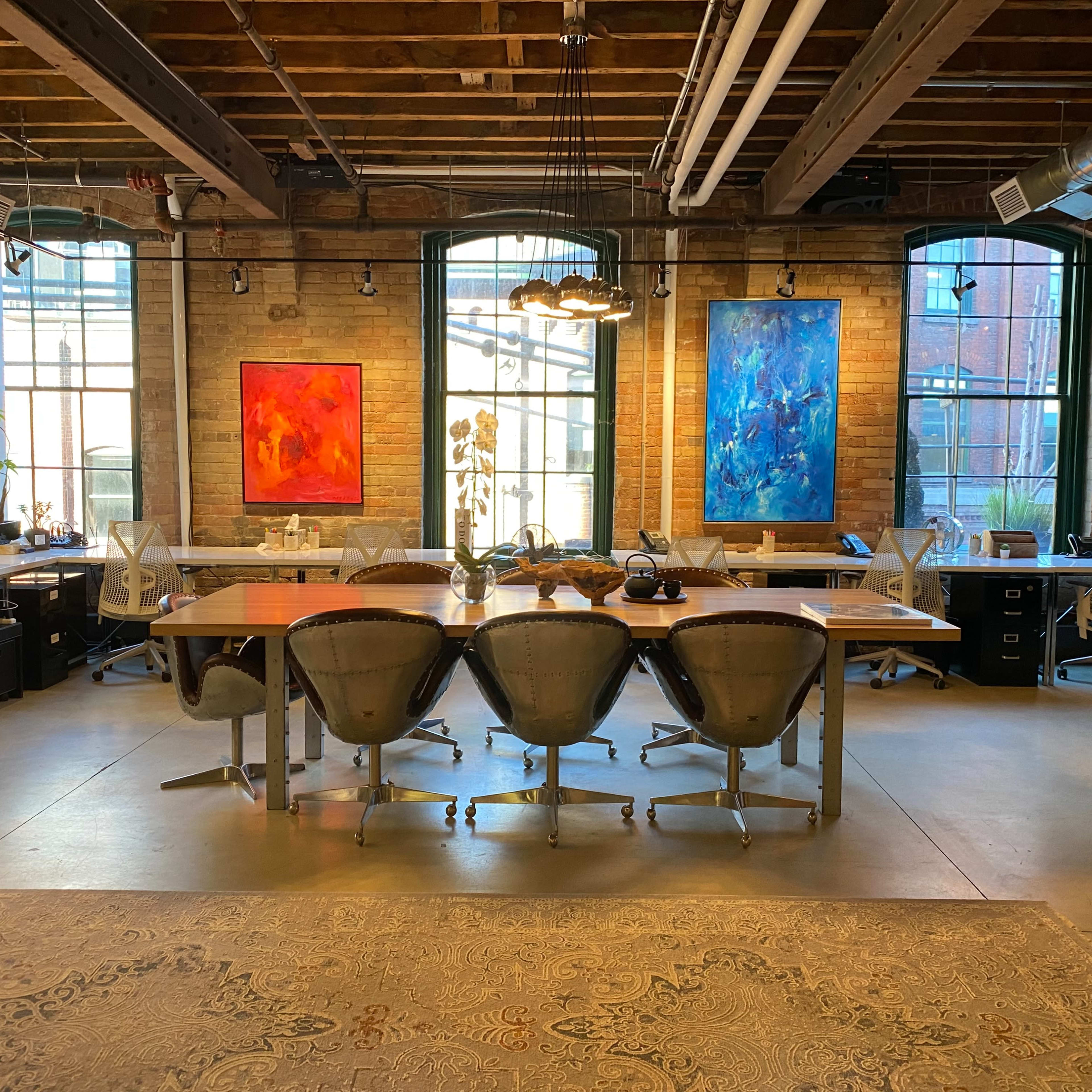A spacious office with large windows, featuring a long wooden table surrounded by modern chairs and colorful abstract paintings on the walls.