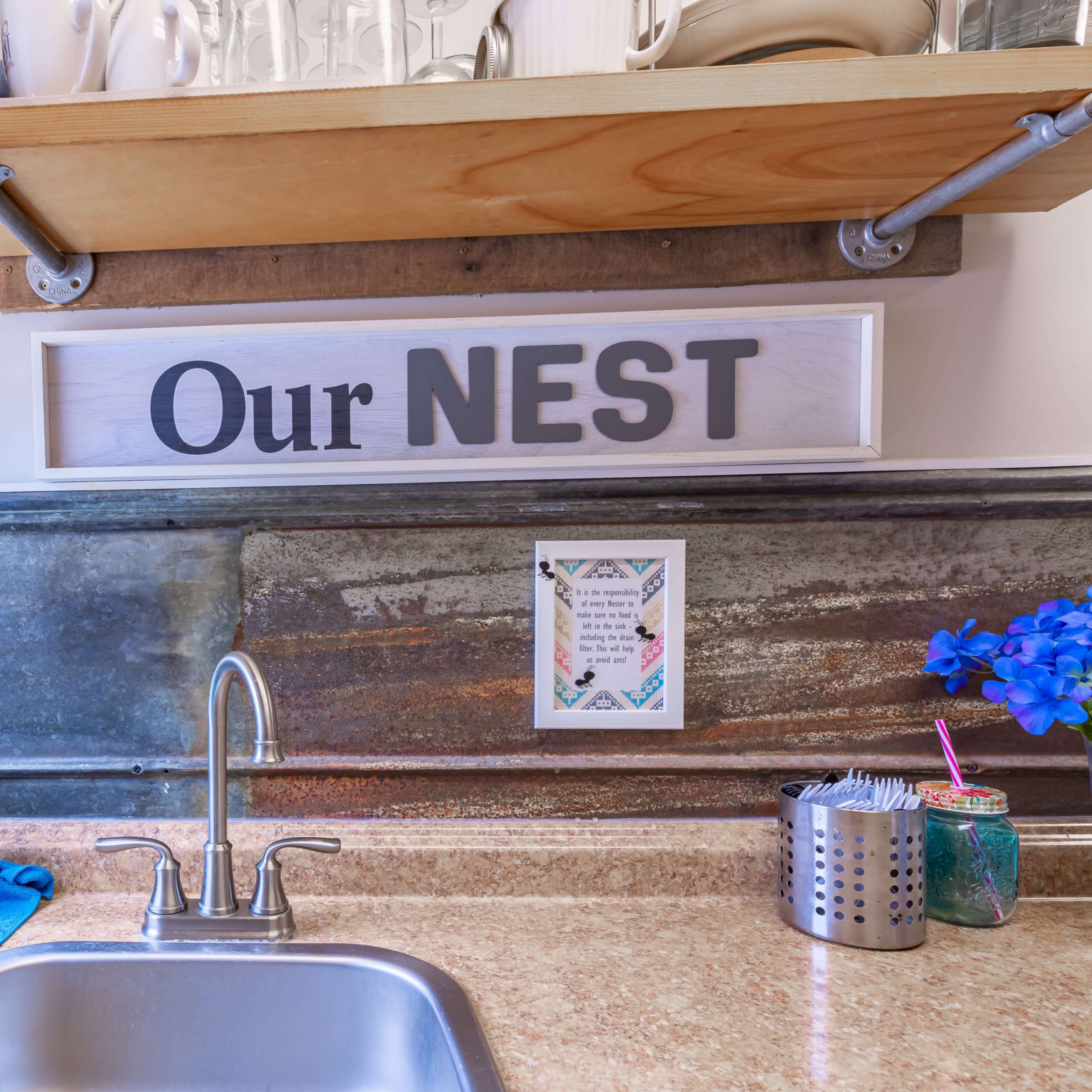 A kitchen sink area is shown with a wooden shelf above it displaying various items, including a sign that reads "Our NEST."