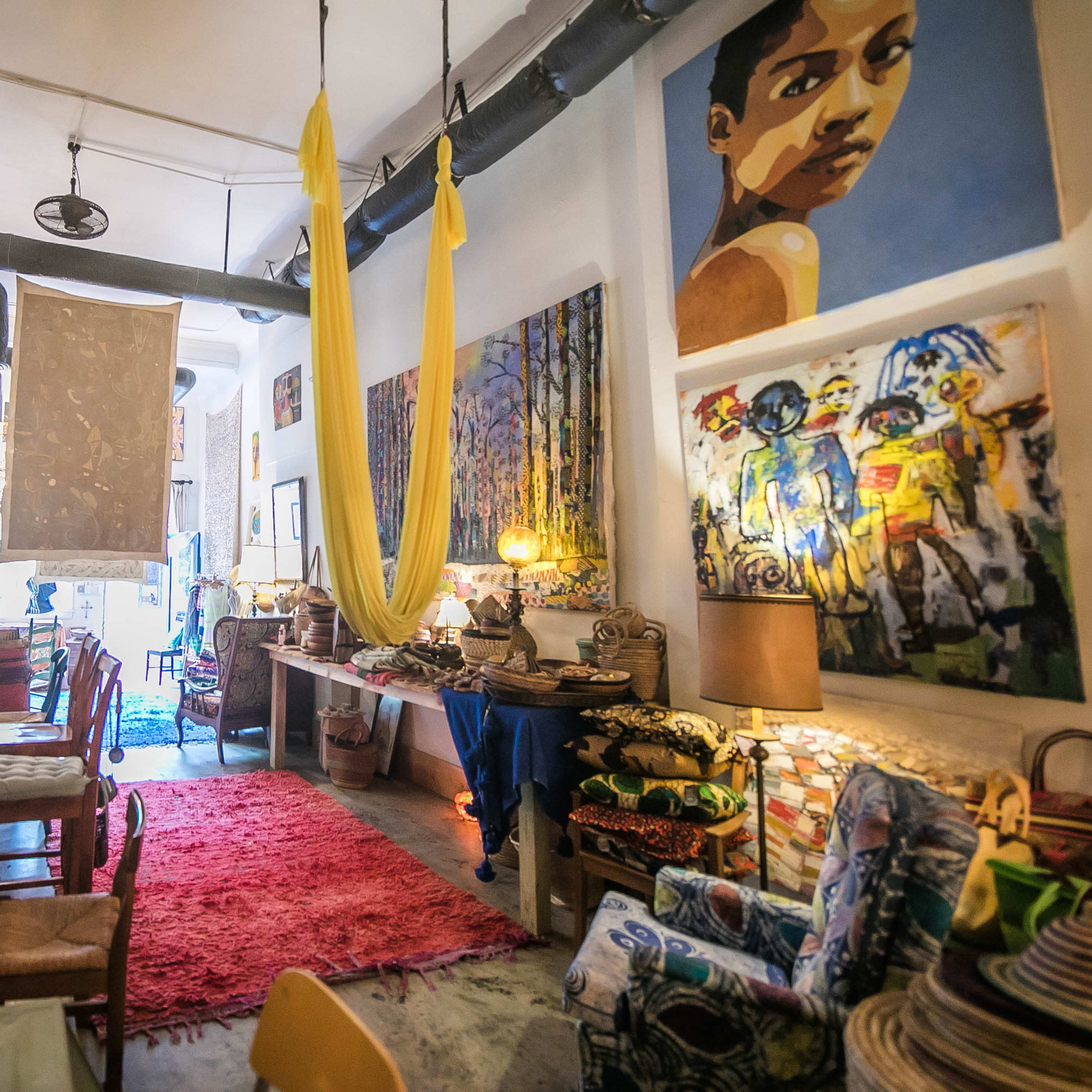 An eclectic art-filled interior of a cozy cafe or shop, featuring colorful paintings, textiles, and various decorative items arranged throughout the space.