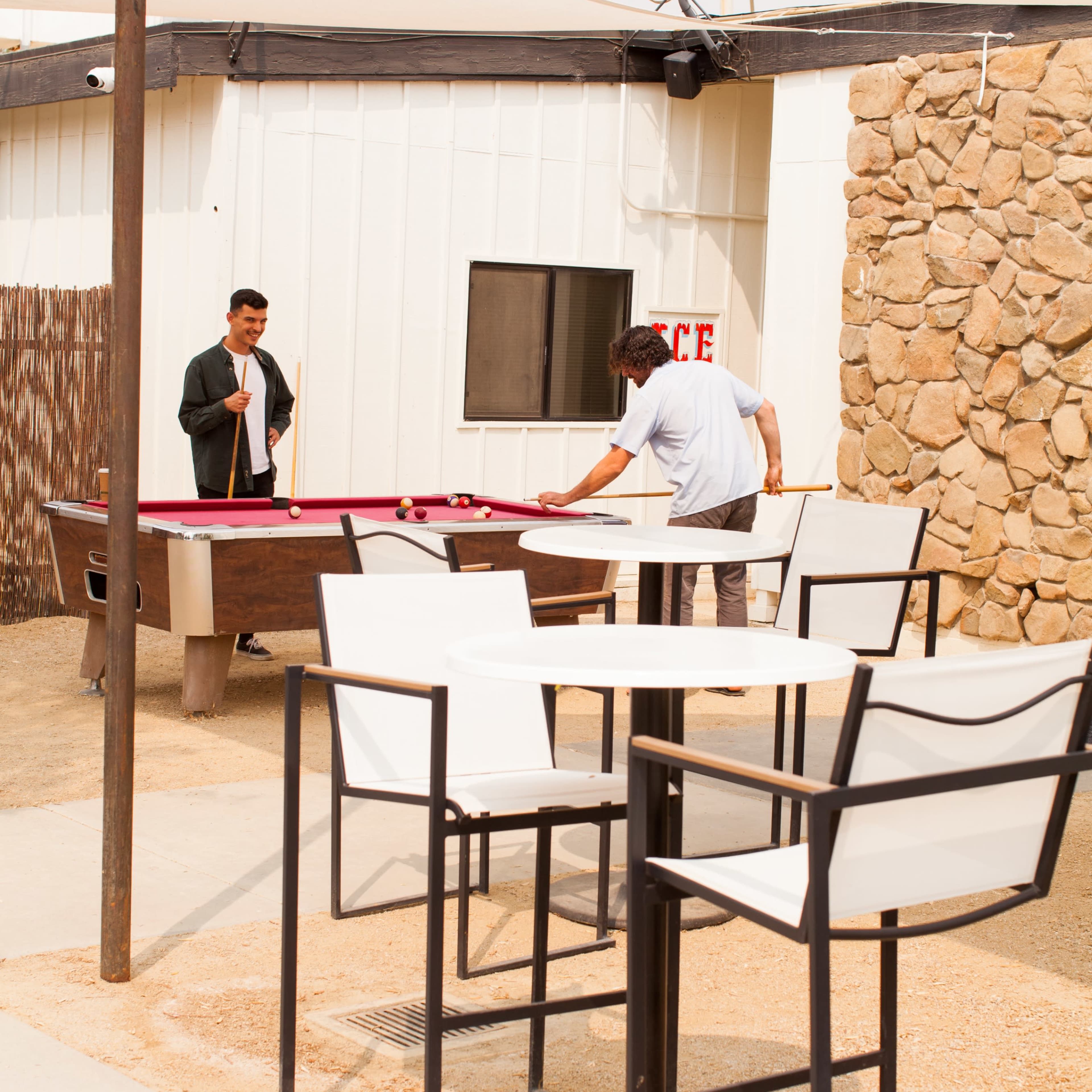 Two men are engaged in a game of pool at an outdoor seating area with a stone wall and a table set nearby.