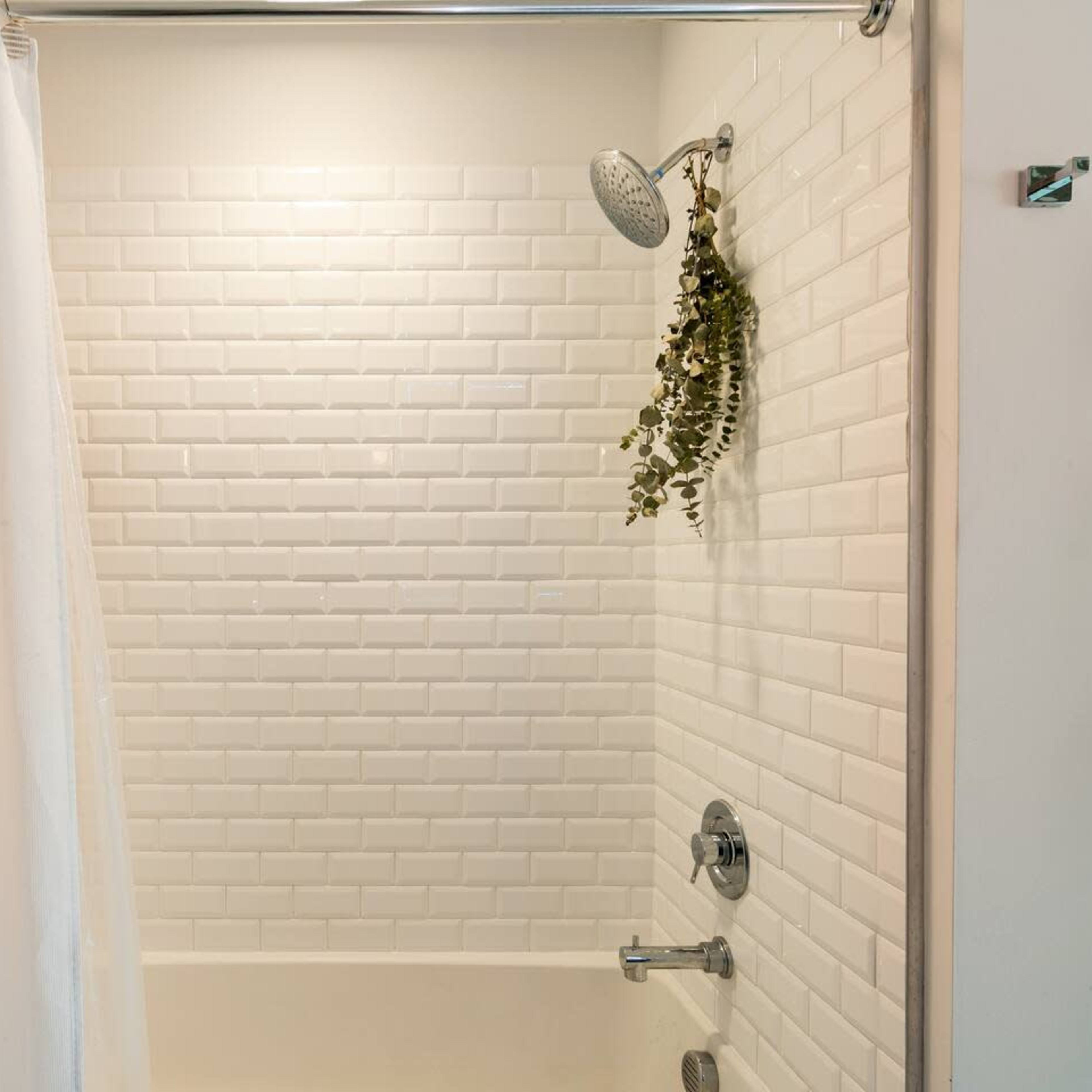 A clean shower area with white subway tiles, a showerhead, and a hanging plant next to a smooth tub.
