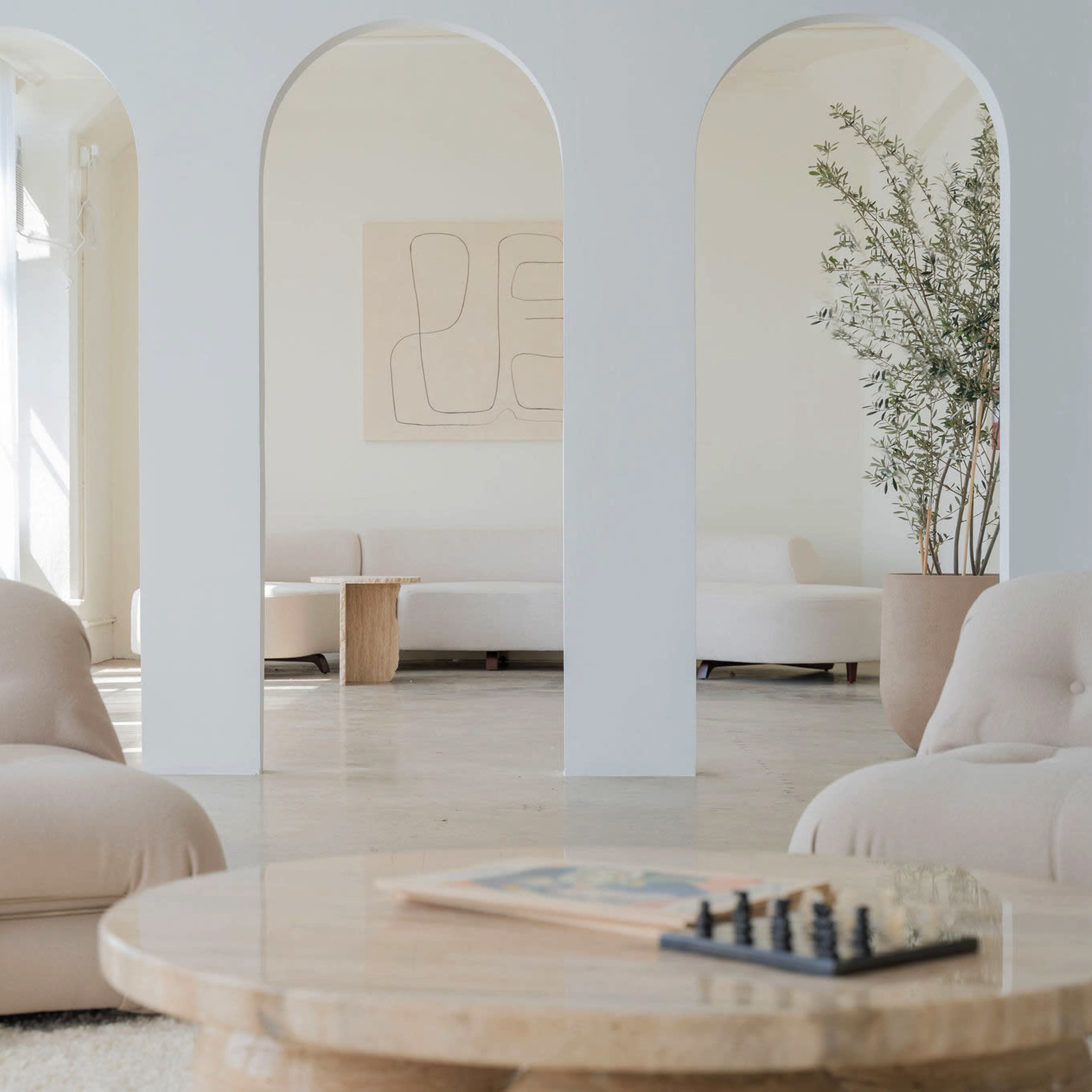 A spacious, minimalist interior with two plush chairs, a round coffee table, and large archways leading to another area with a sofa and a potted plant.