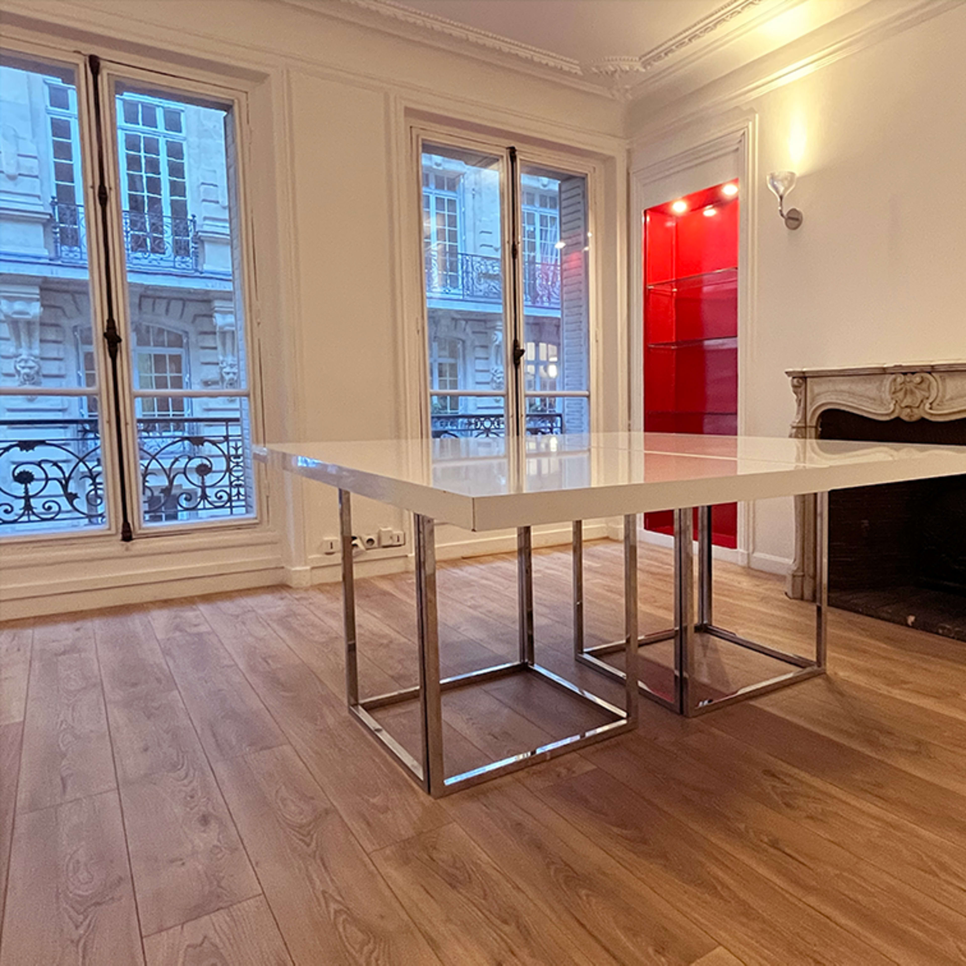 A modern room featuring a white square table with chrome legs, positioned near a fireplace and large windows.