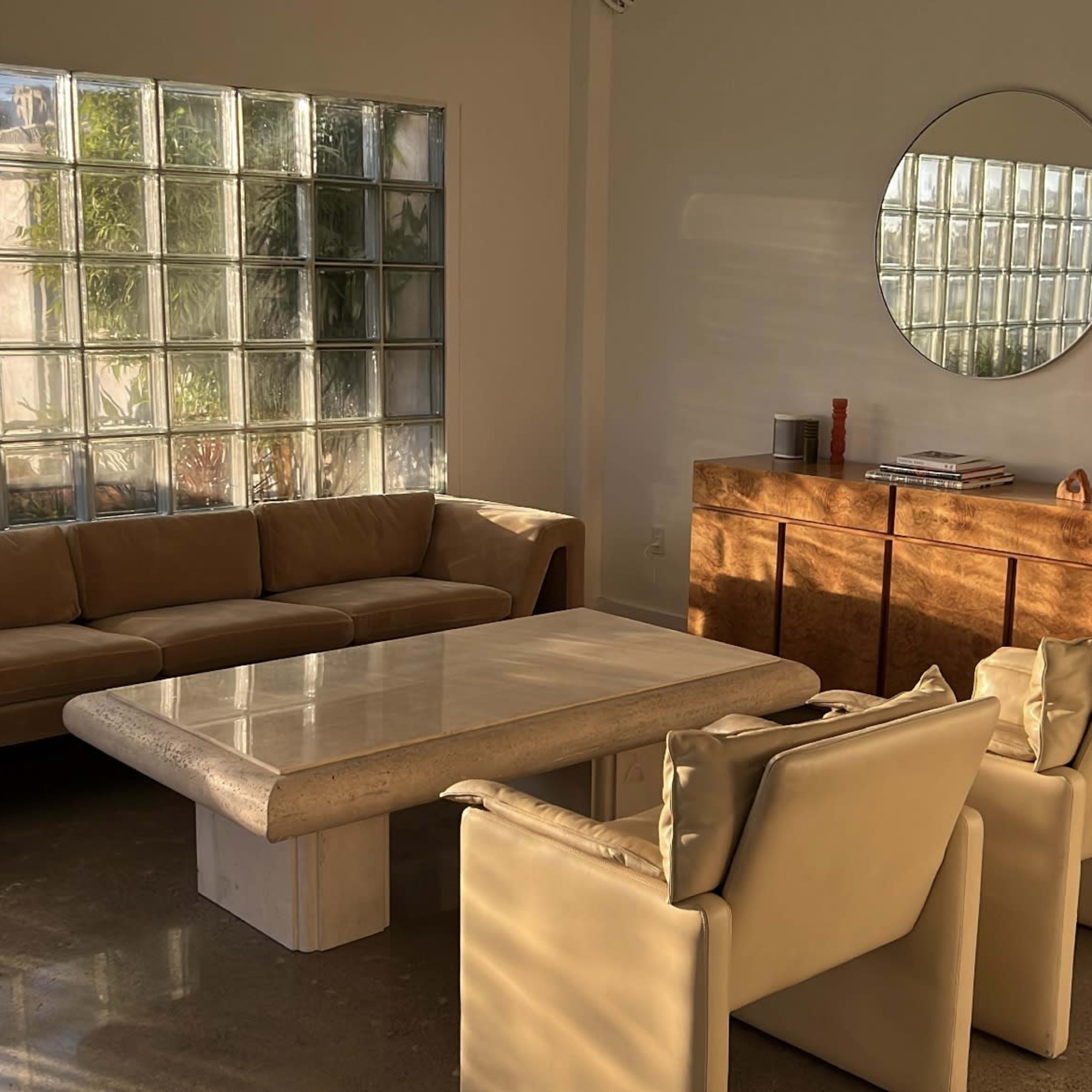 A modern living room with a large beige sofa, a stone coffee table, two light-colored armchairs, and a wooden sideboard, illuminated by natural light from a glass block wall.