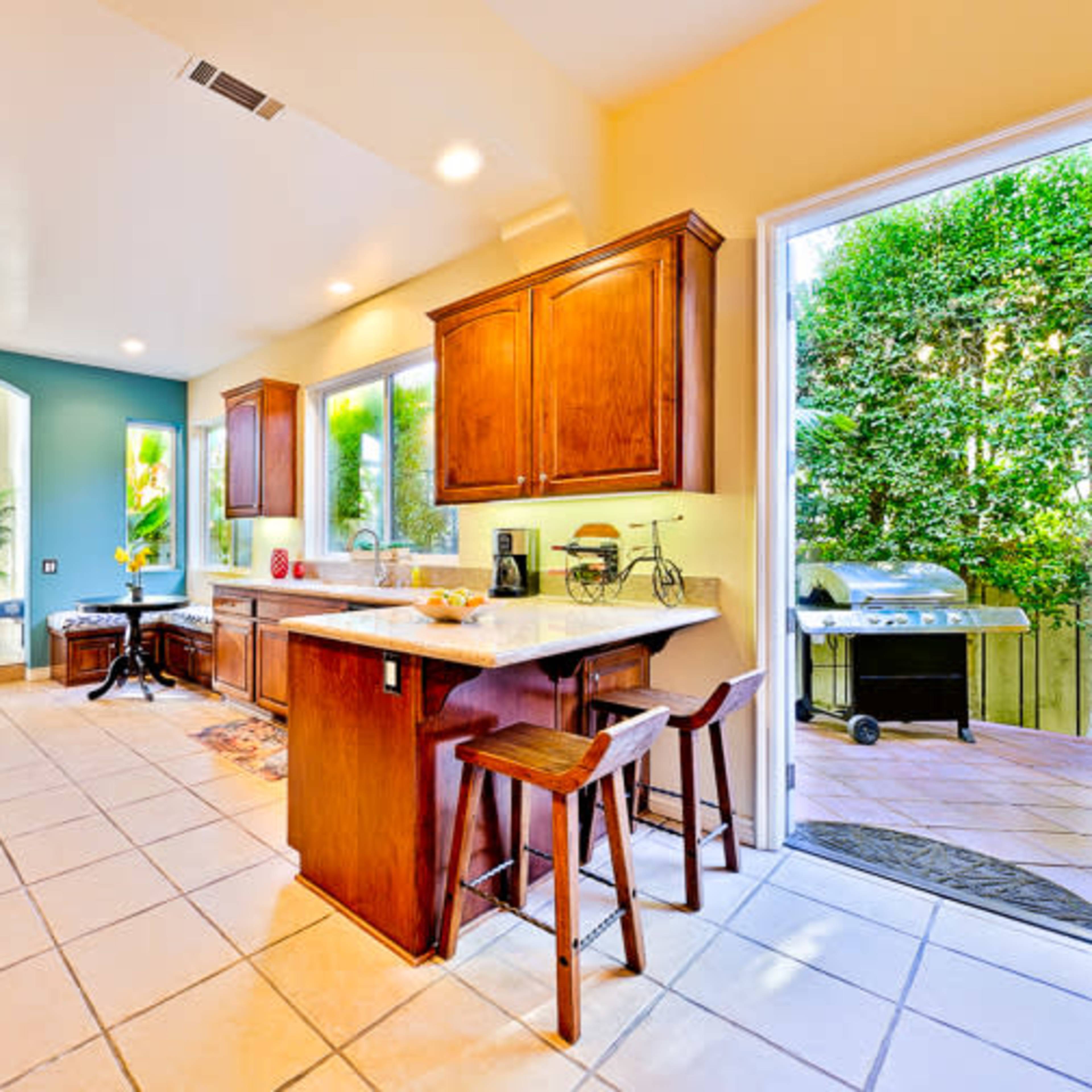 A kitchen features wooden cabinetry, a central island with bar stools, and large windows that overlook a patio with outdoor seating and a grill.