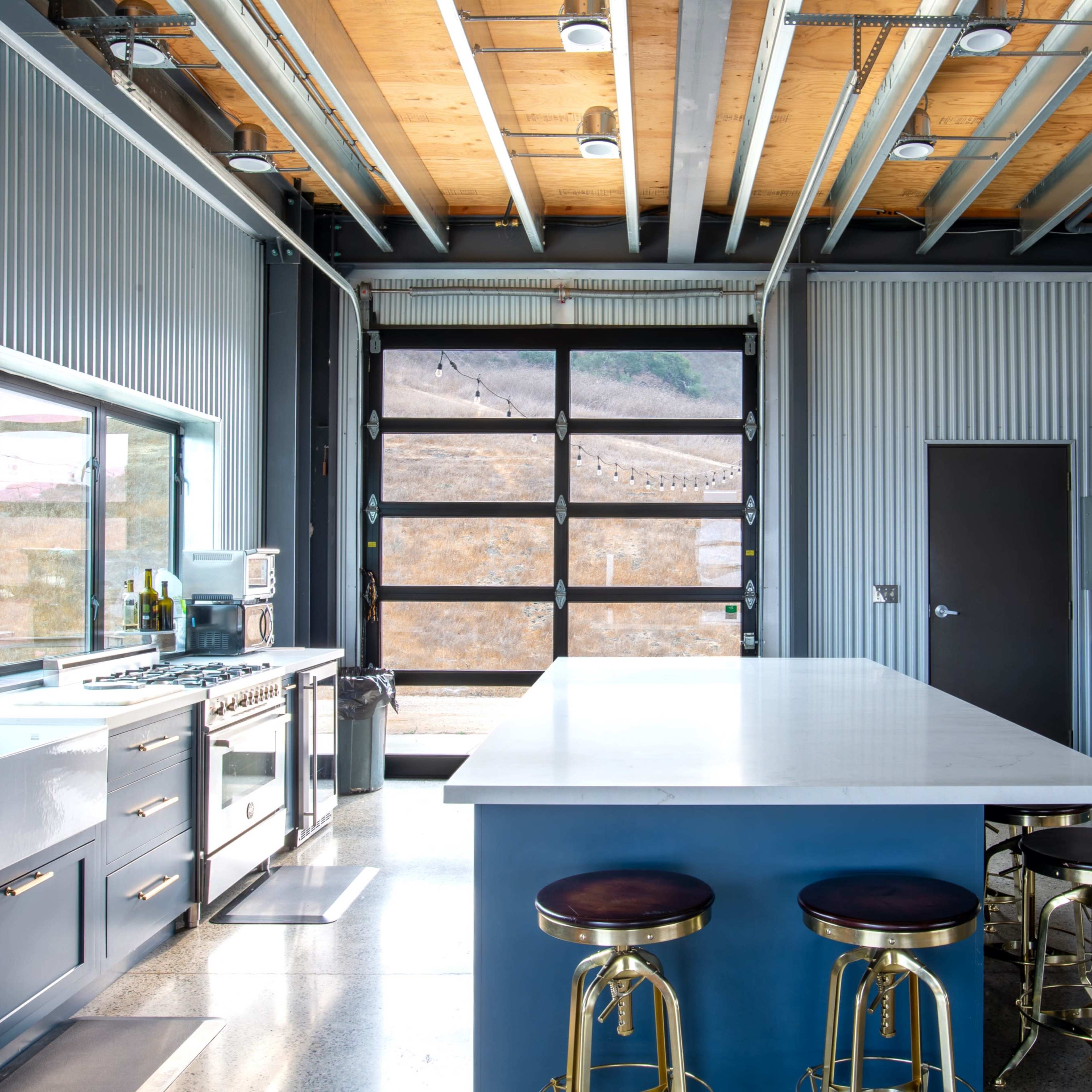 The kitchen features a large island with bar stools, blue cabinetry, stainless steel appliances, and a wall of windows overlooking a large garage door.