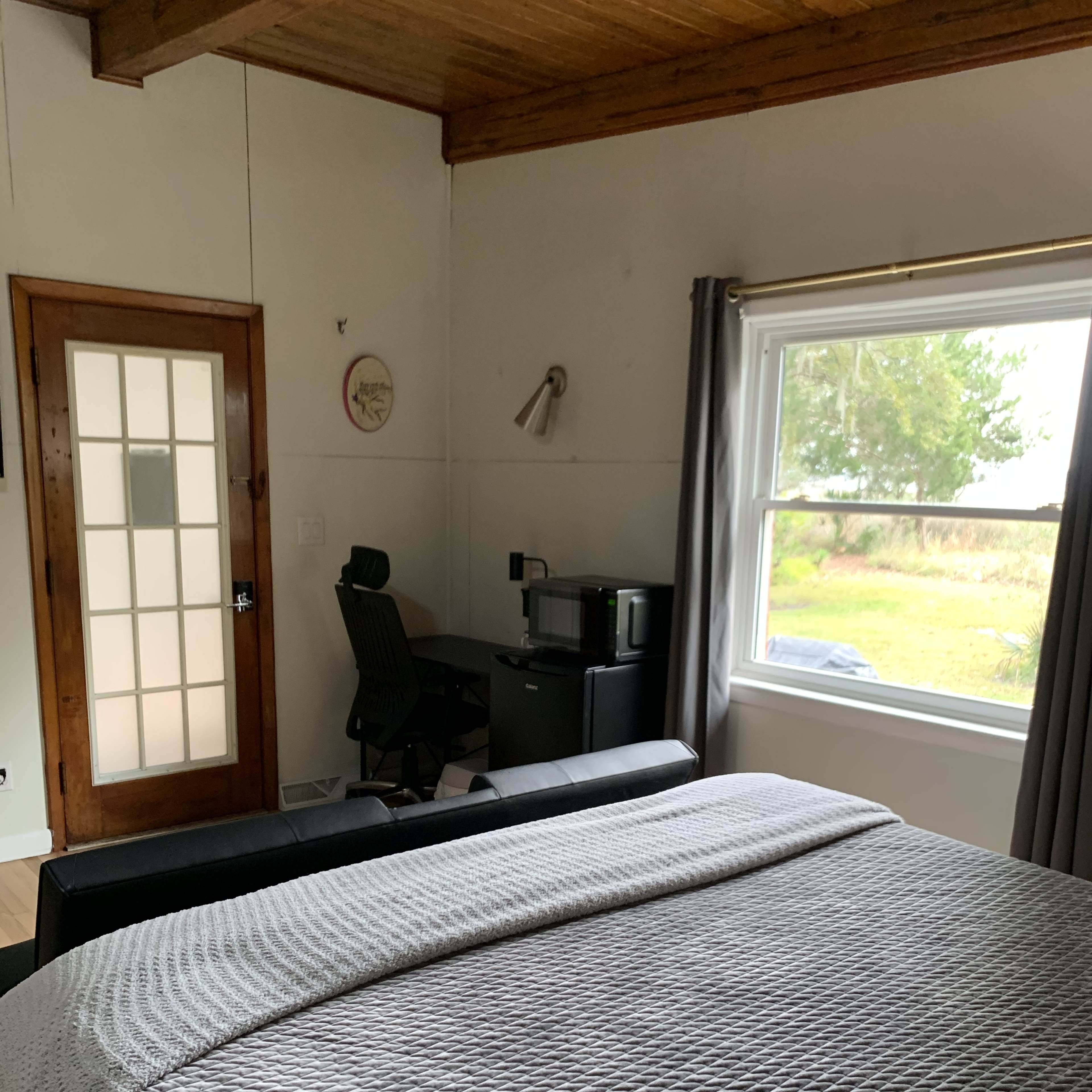 A bedroom with a bed, a chair, a small desk, and a window overlooking a grassy area.