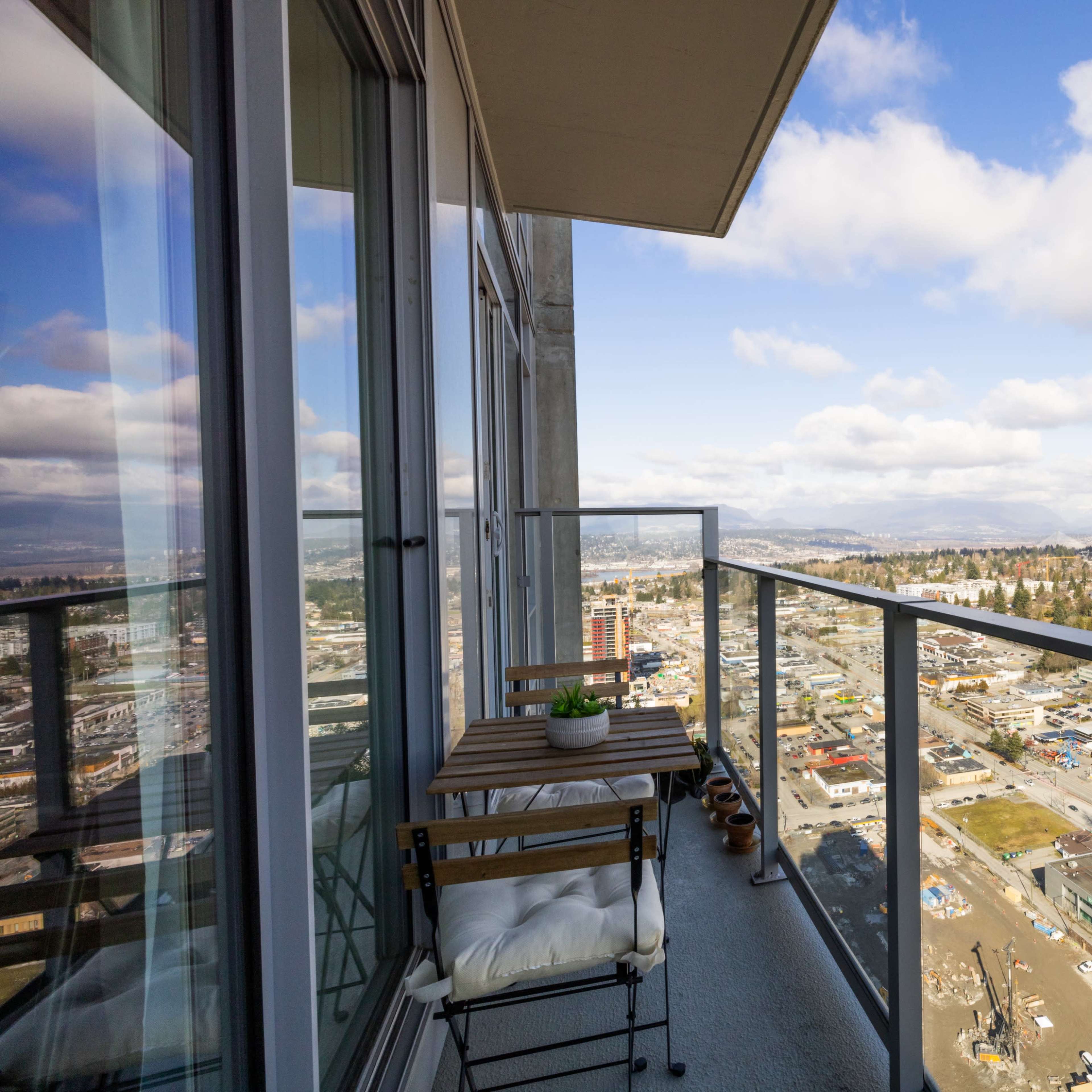 A balcony with a small table and chair overlooks a cityscape featuring buildings and roads under a partly cloudy sky.