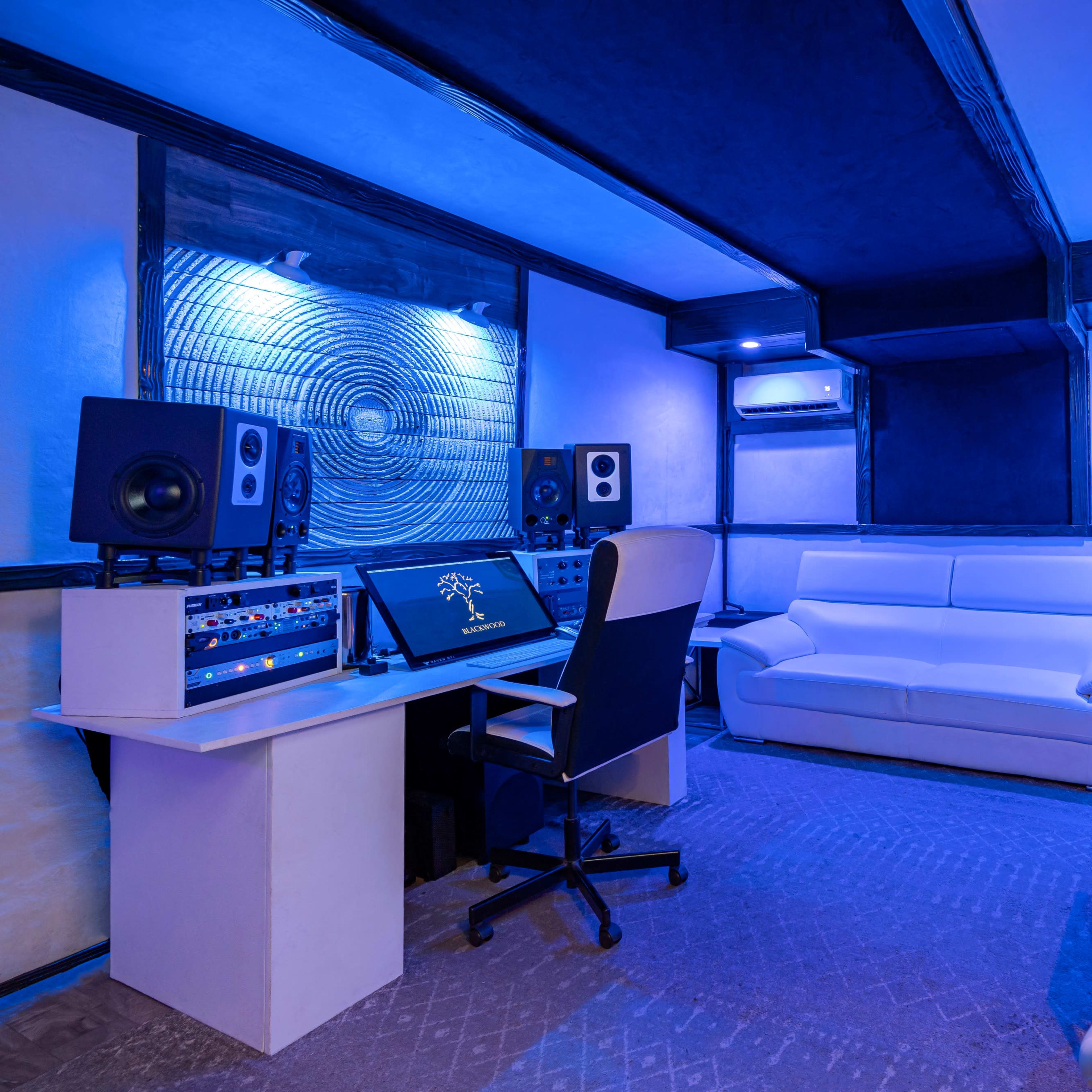 A music studio features a control desk with speakers, a white sofa, and modern seating, all illuminated by blue lighting.