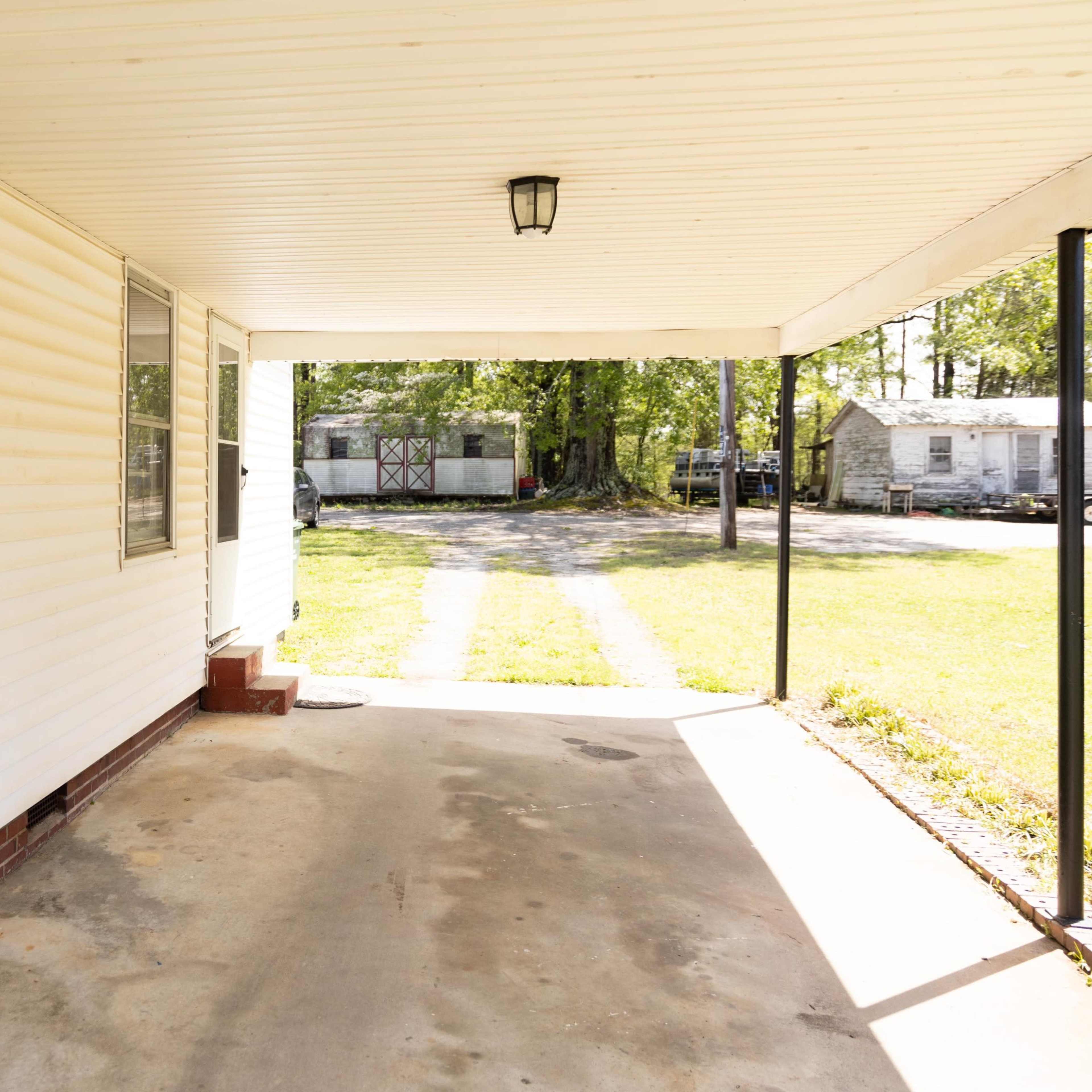 A covered carport attached to a house, with a concrete floor and a view of an open yard and additional structures in the background.