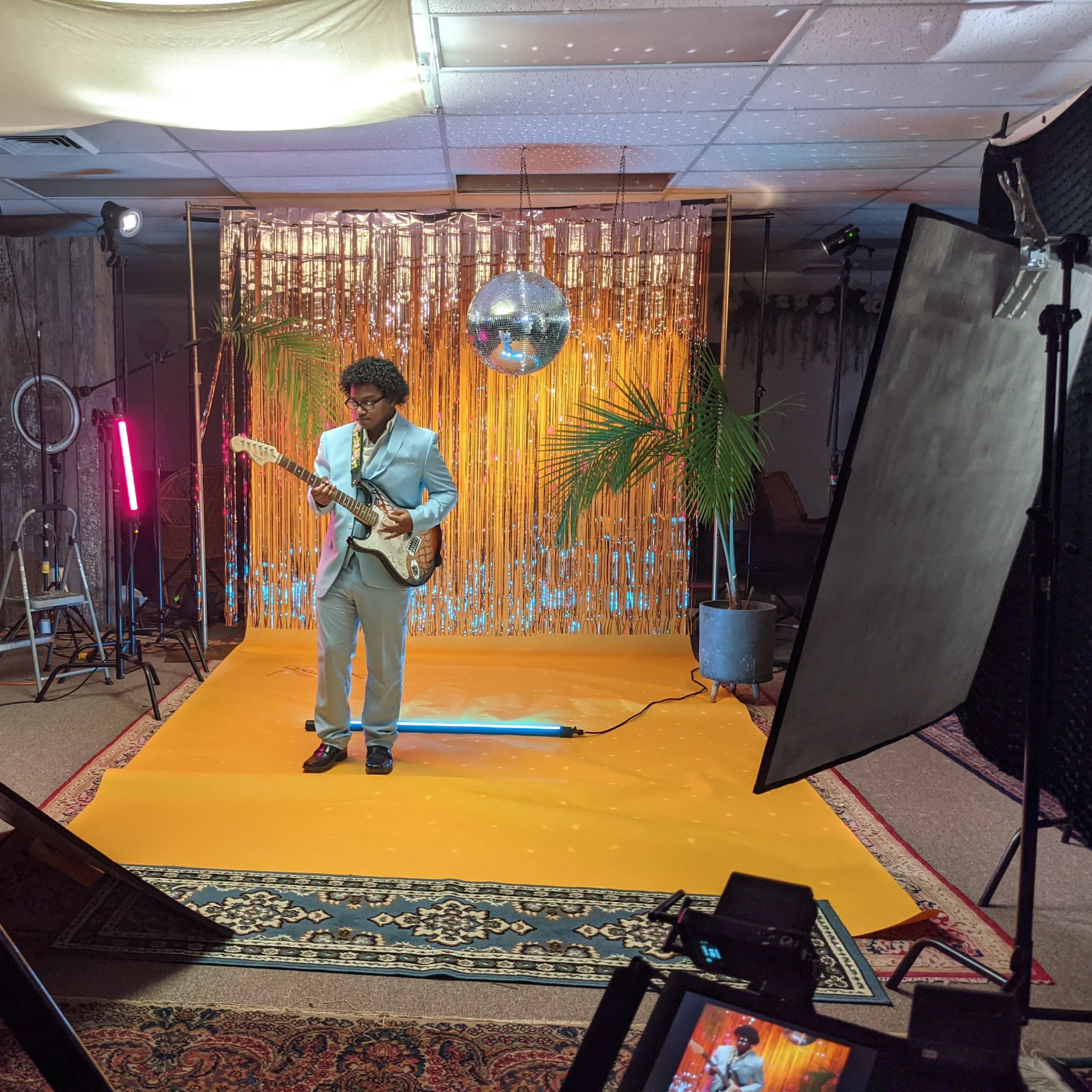A musician in a light blue suit plays an electric guitar in front of a sequined backdrop and a disco ball, surrounded by studio lighting and plants.