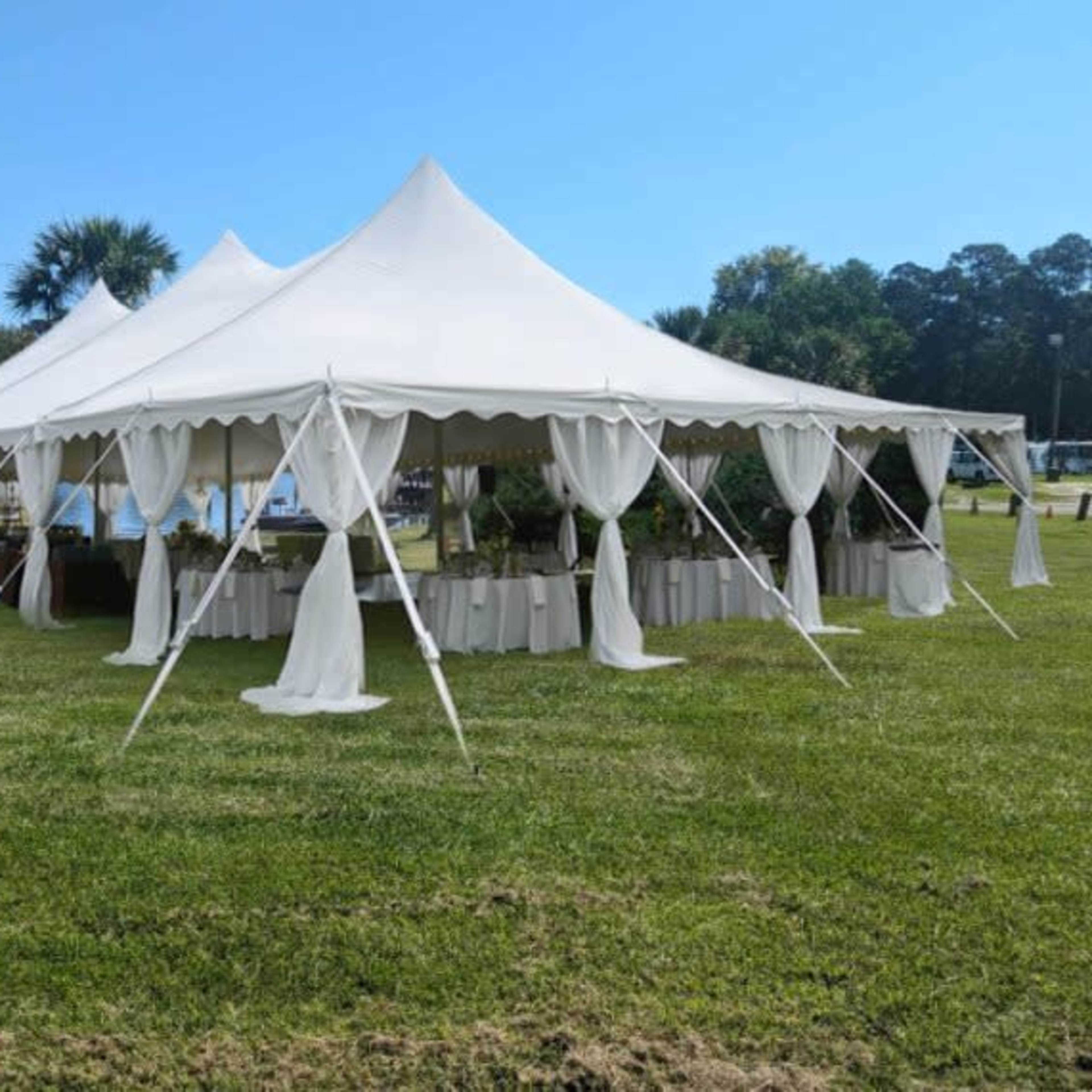 A large white tent with draped sides is set up on a grassy area beside a lake.