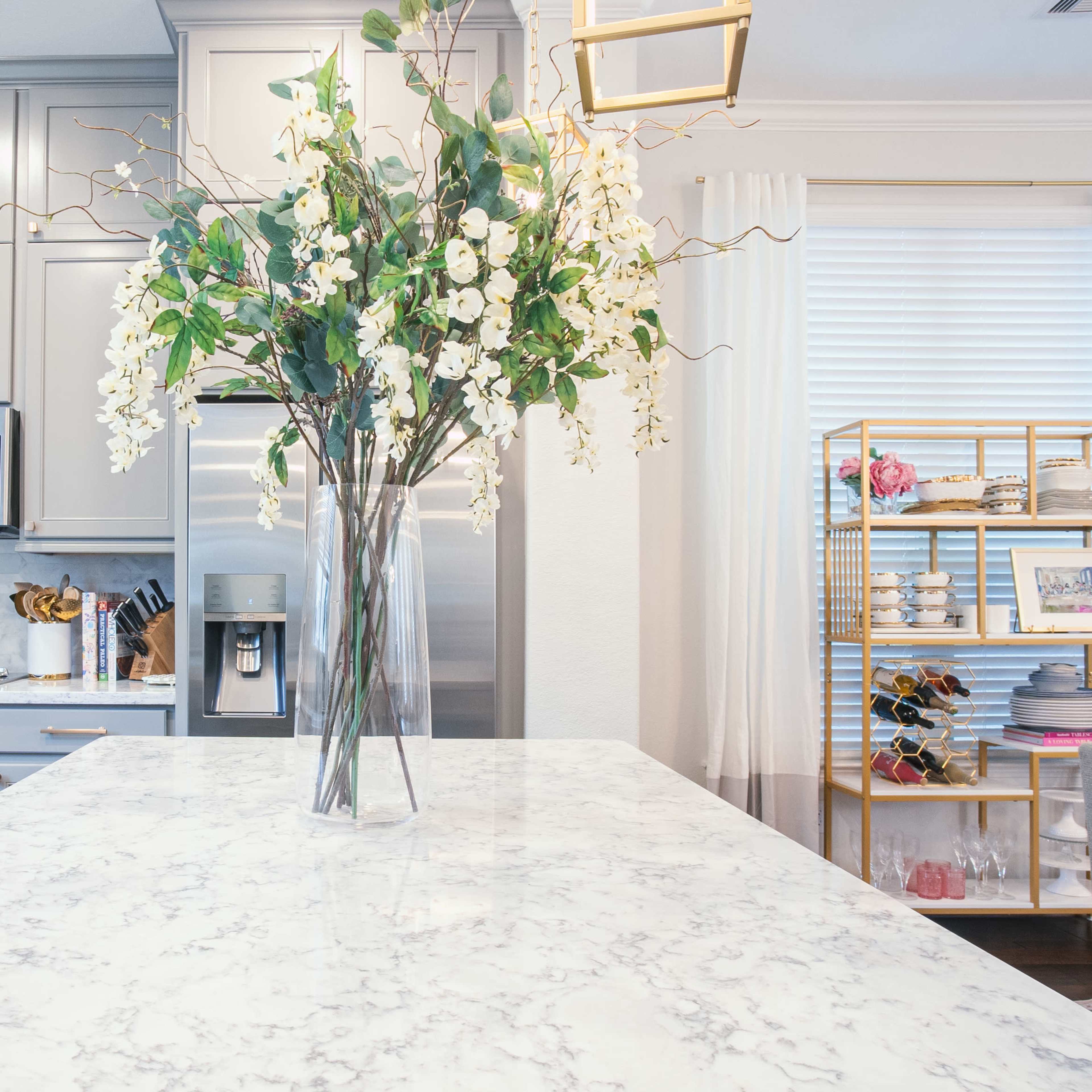 A bright kitchen features gray cabinetry, stainless steel appliances, and a marble island with a large vase of flowers.