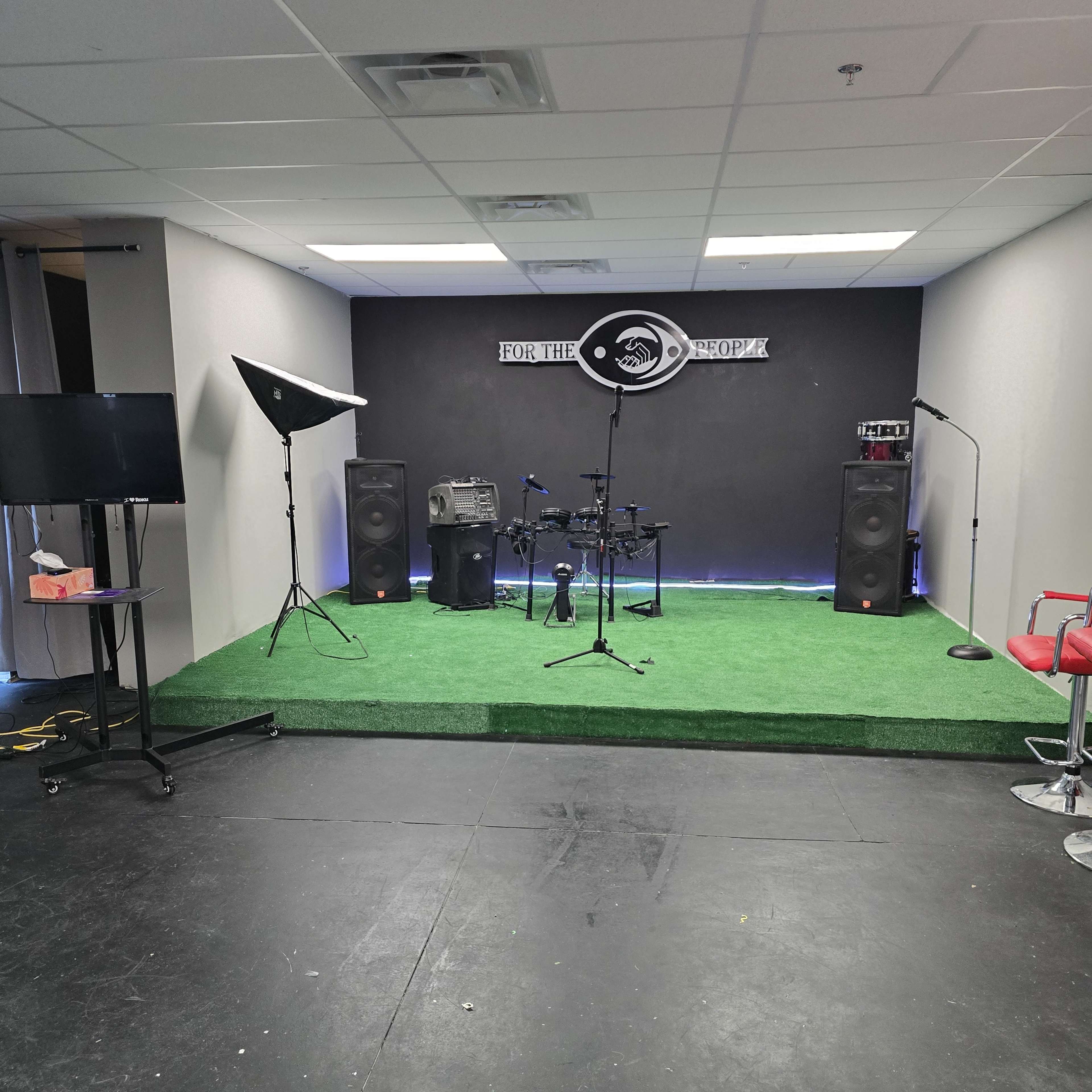 A small stage area with musical equipment, including a drum set and speakers, set against a black wall and green carpeting.