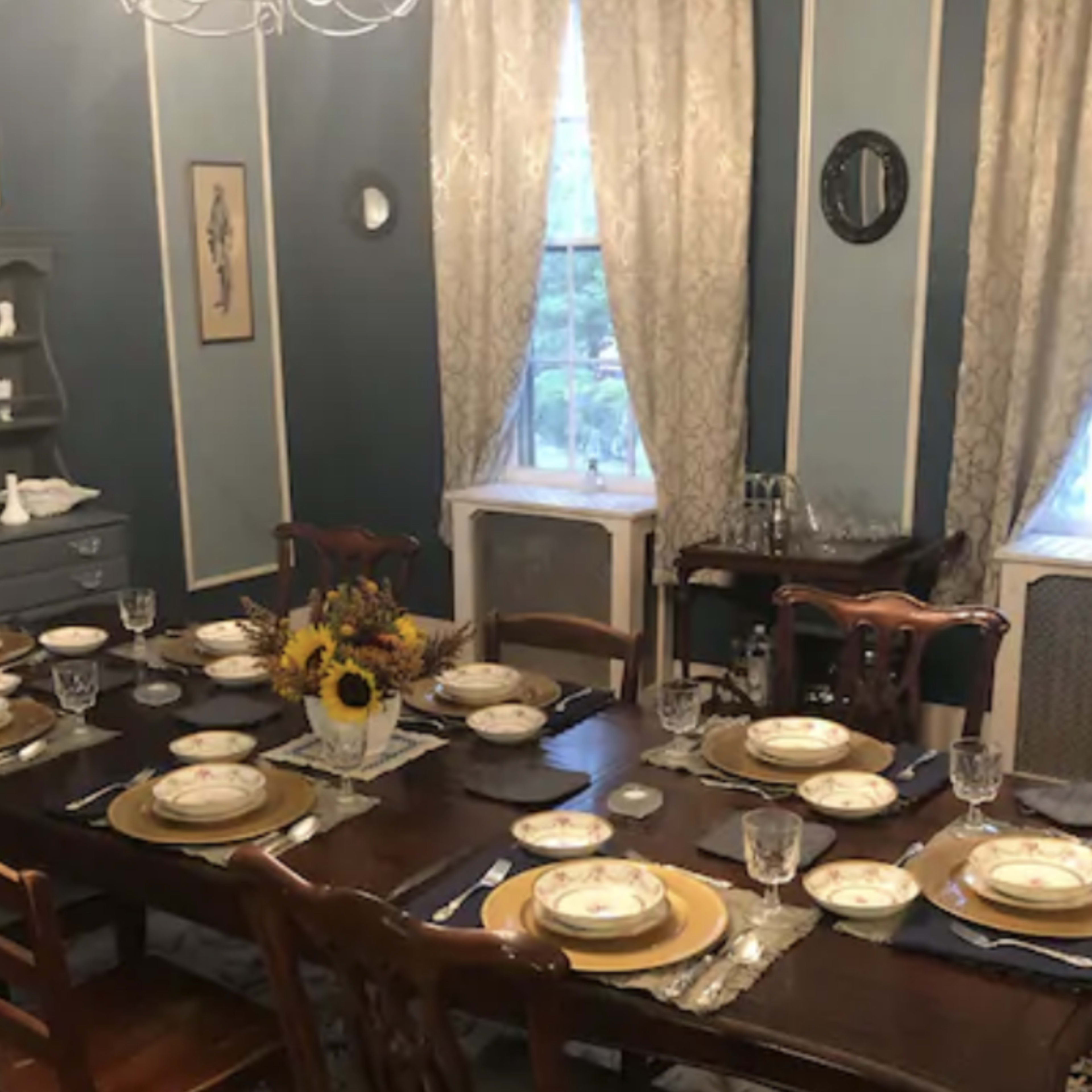 A formal dining room is set with a large wooden table surrounded by chairs, adorned with plates, glasses, and a vase of flowers, while blue and white walls frame the scene.