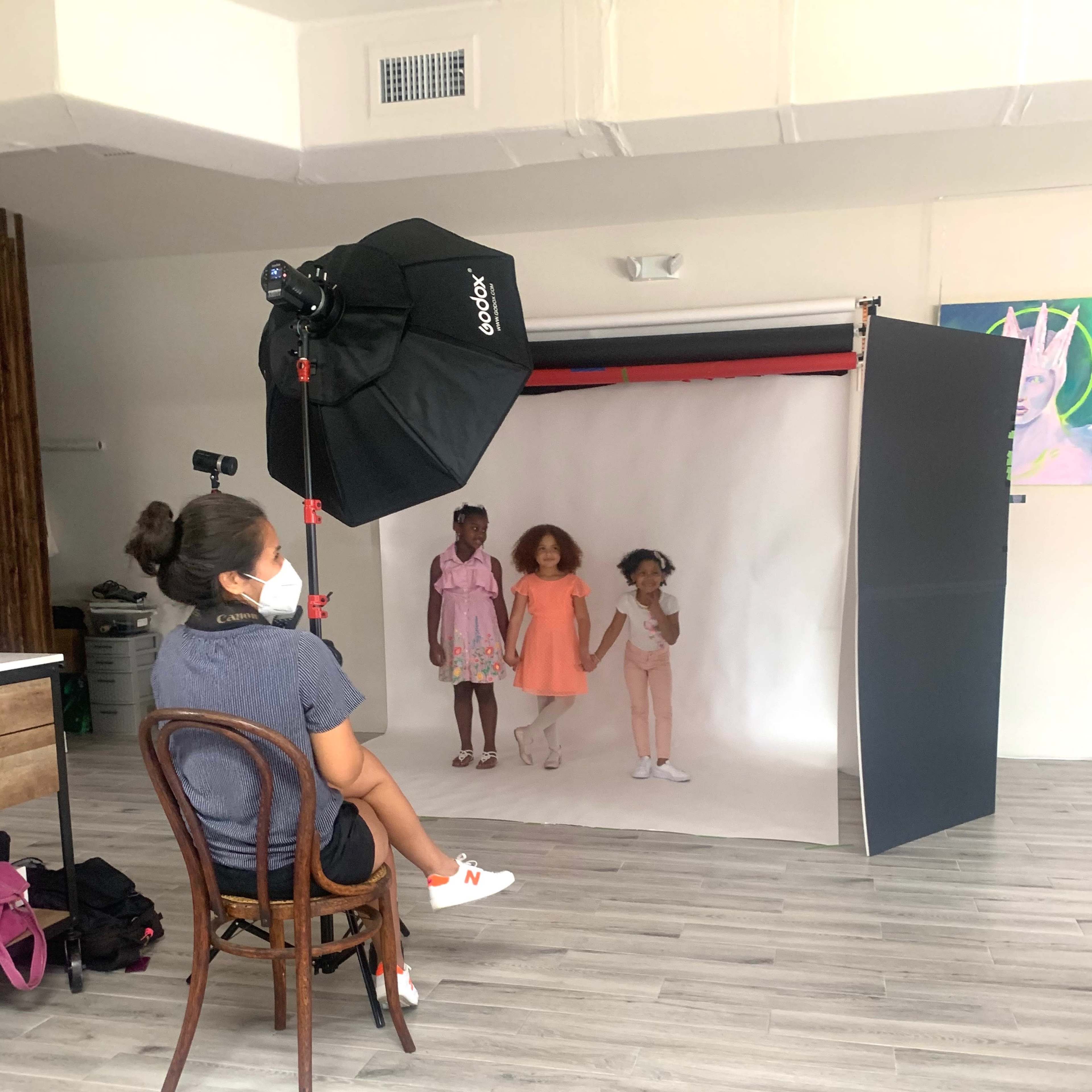 A photographer sits on a chair in a studio while three children pose together in front of a backdrop.
