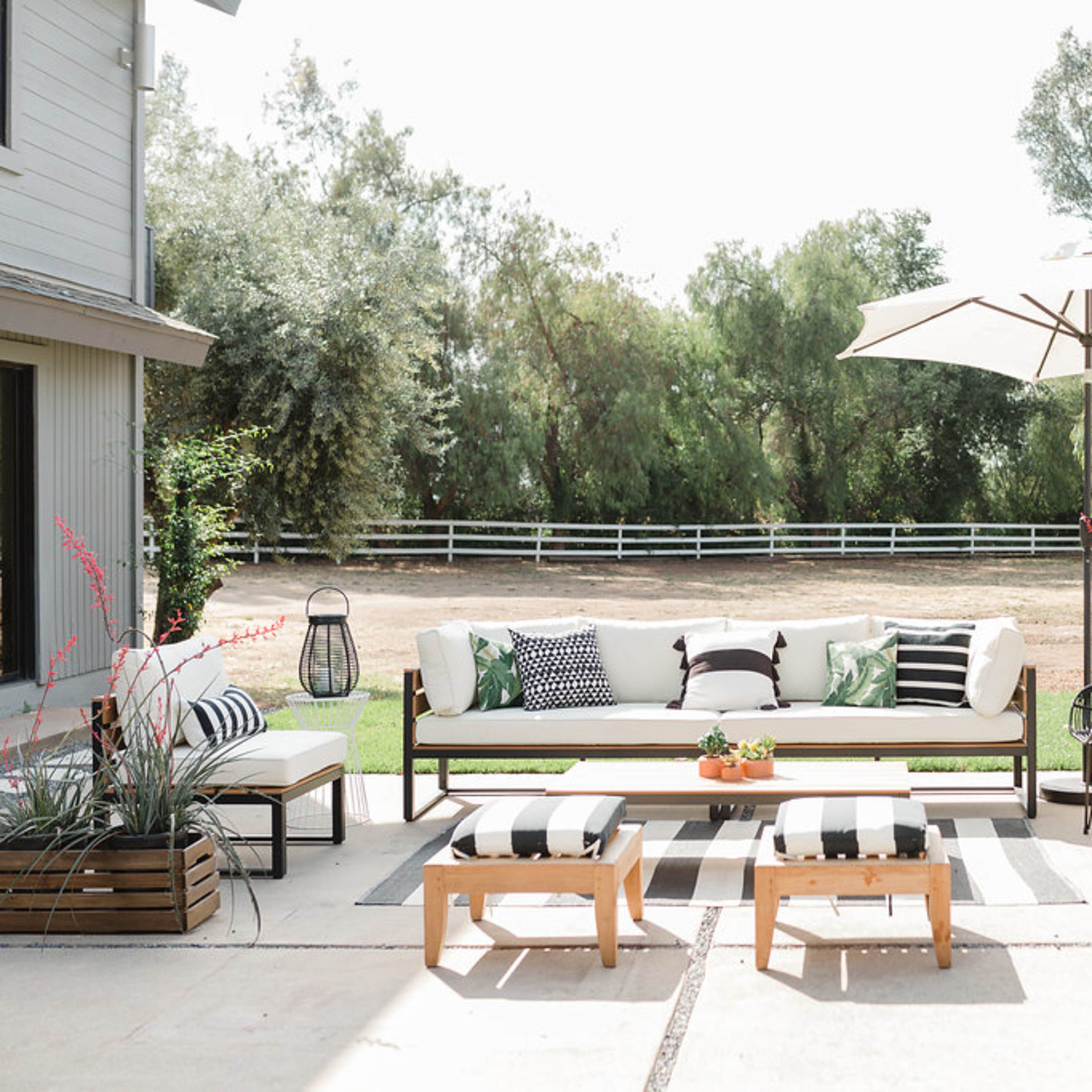 A stylish outdoor seating area with a sofa, chairs, and an umbrella setup on a patio, surrounded by greenery and a fenced yard.