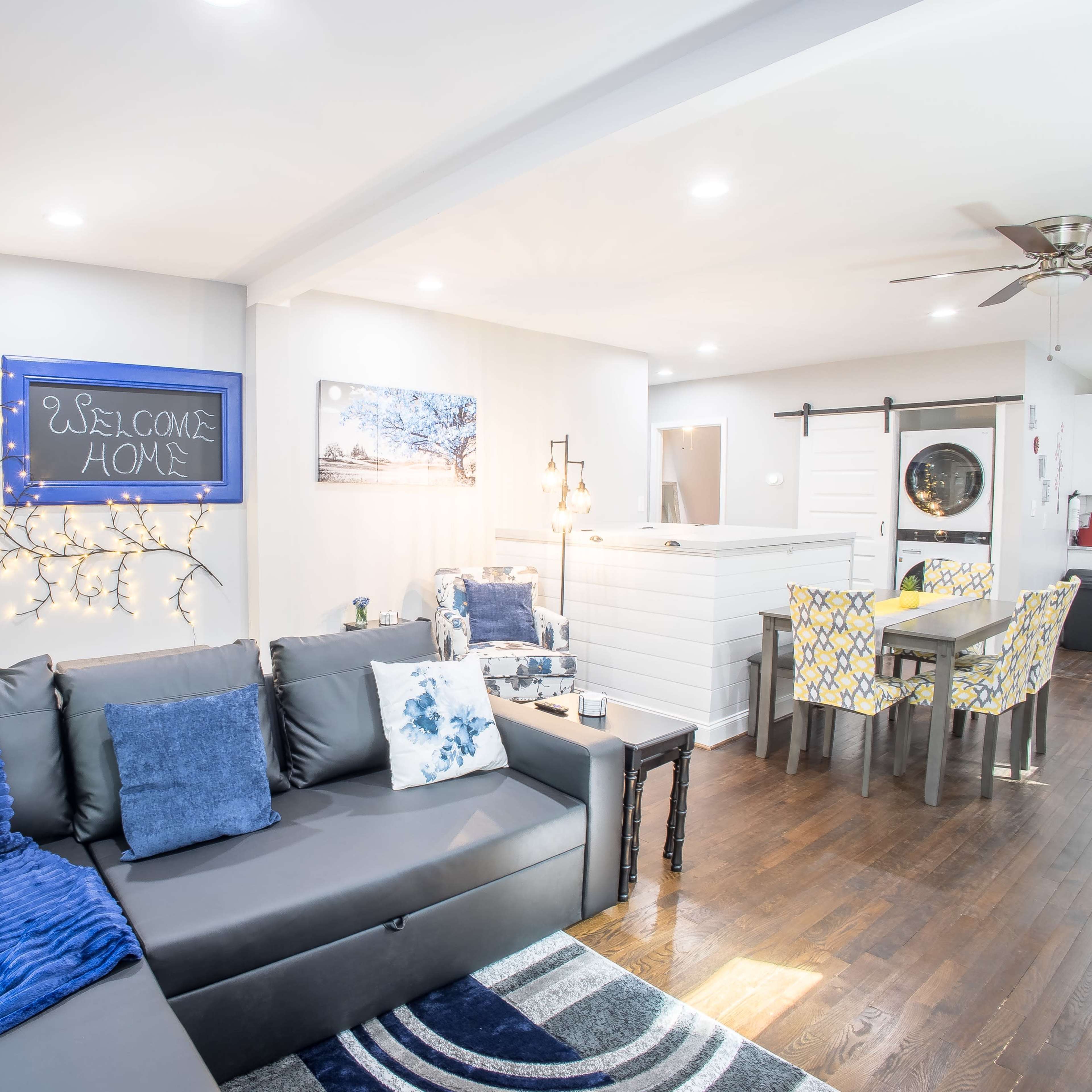 A modern living room with a gray sofa, a welcome sign on the wall, and a dining area in the background.