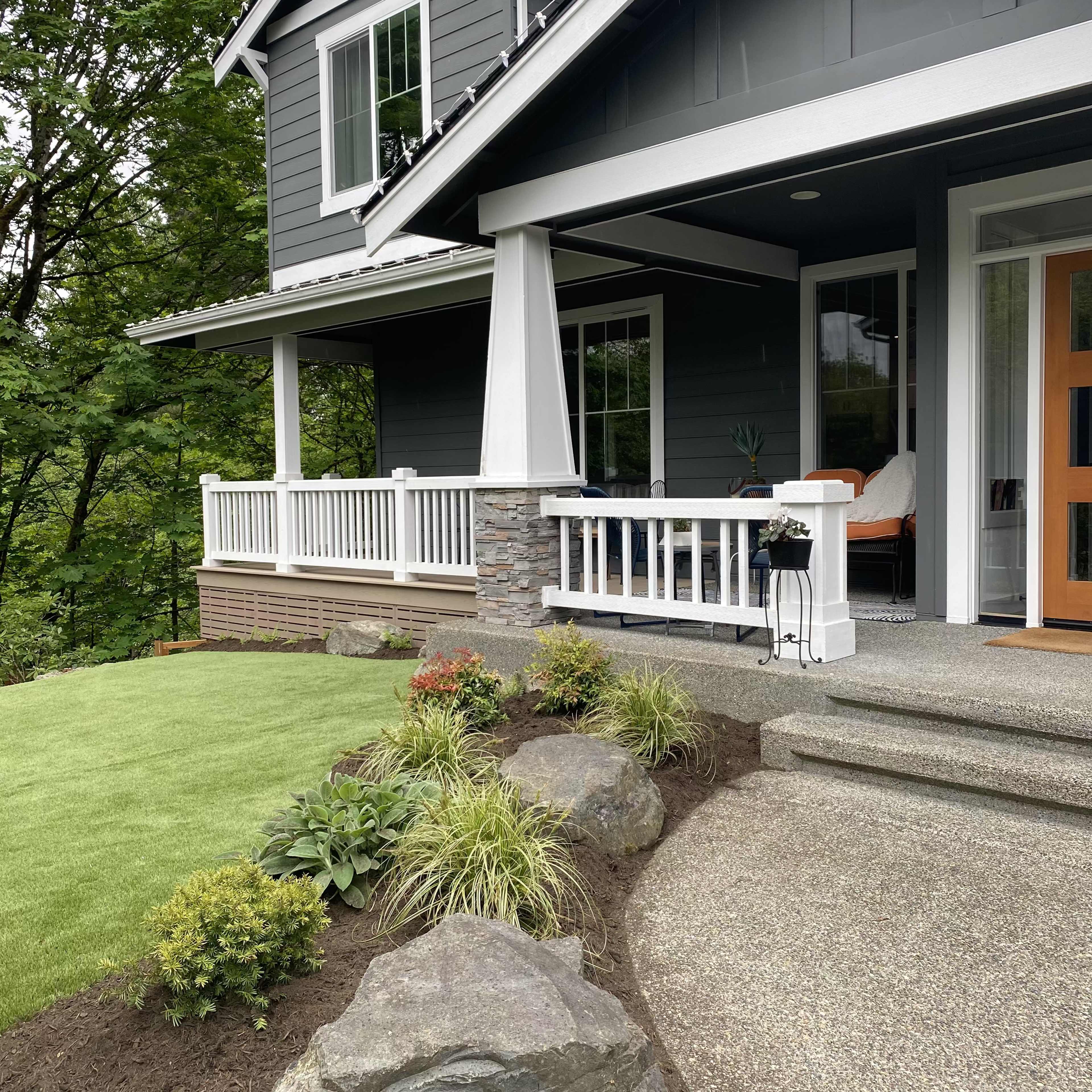 A contemporary house with a dark gray exterior, a covered porch, and landscaped areas featuring plants and rocks.