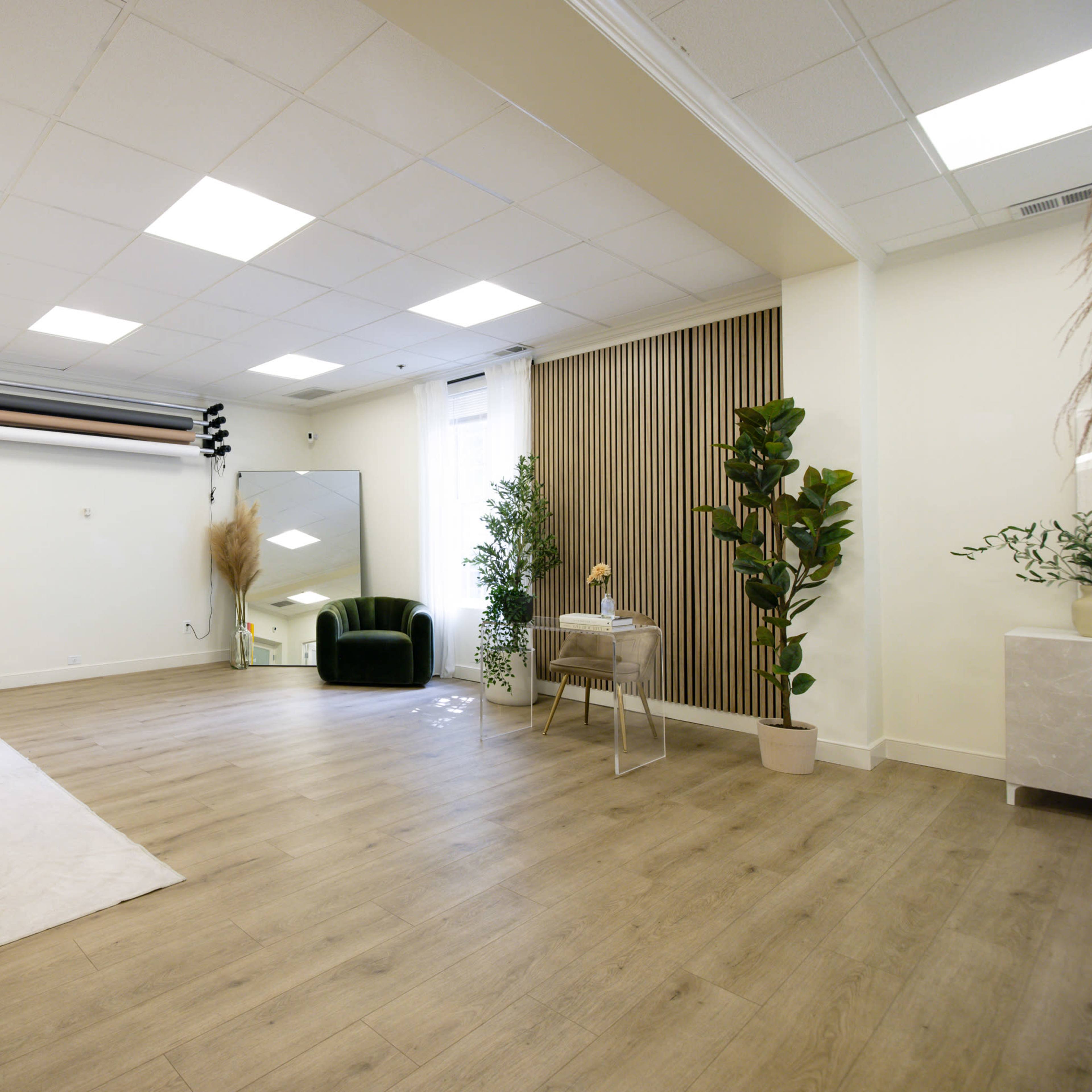 A spacious interior room features a white sofa, a green chair, a small table, and large plants, with ample natural light coming through the windows.