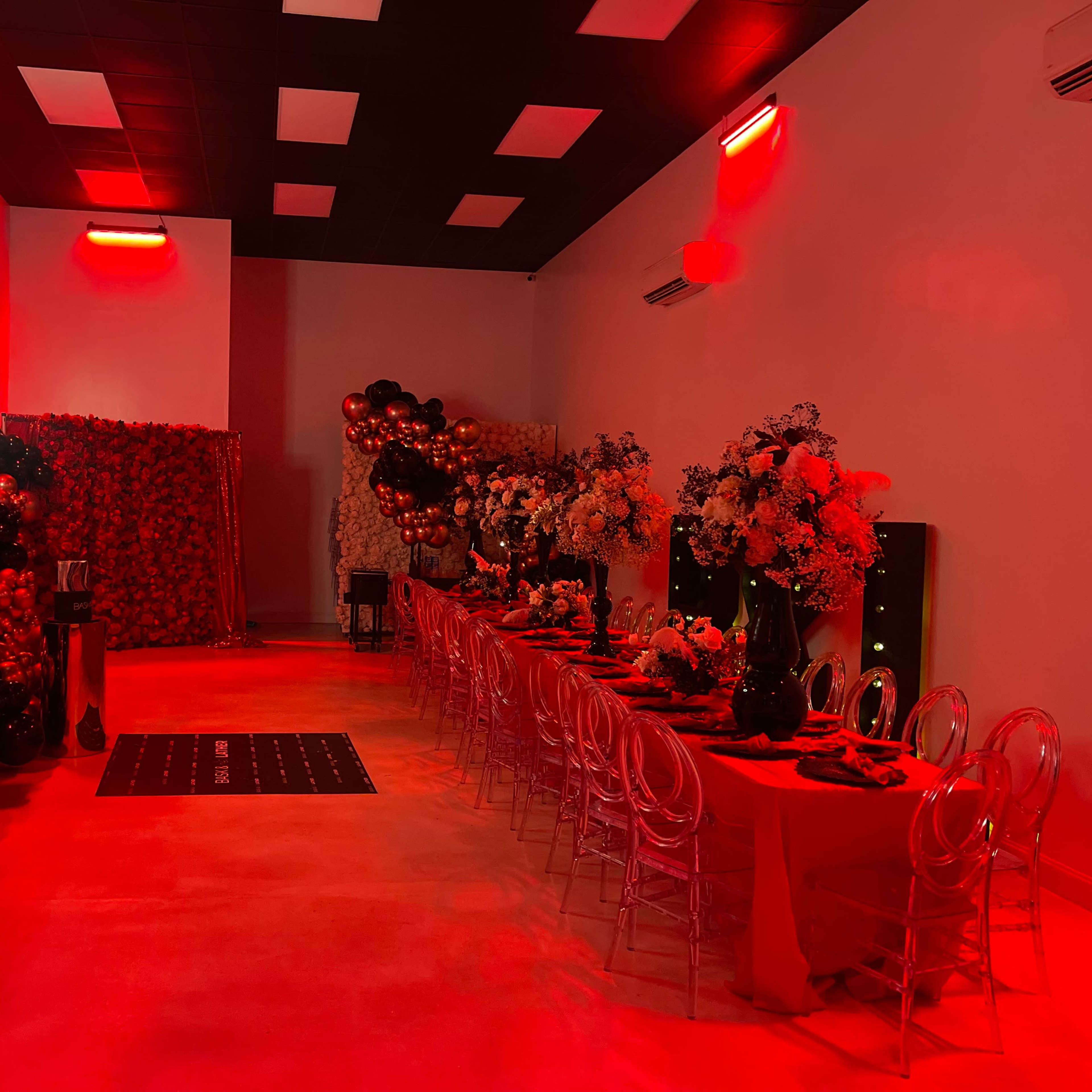 A decorated event space featuring a long dining table set with red tablecloths, surrounded by colorful balloon arrangements and floral centerpieces under red lighting.