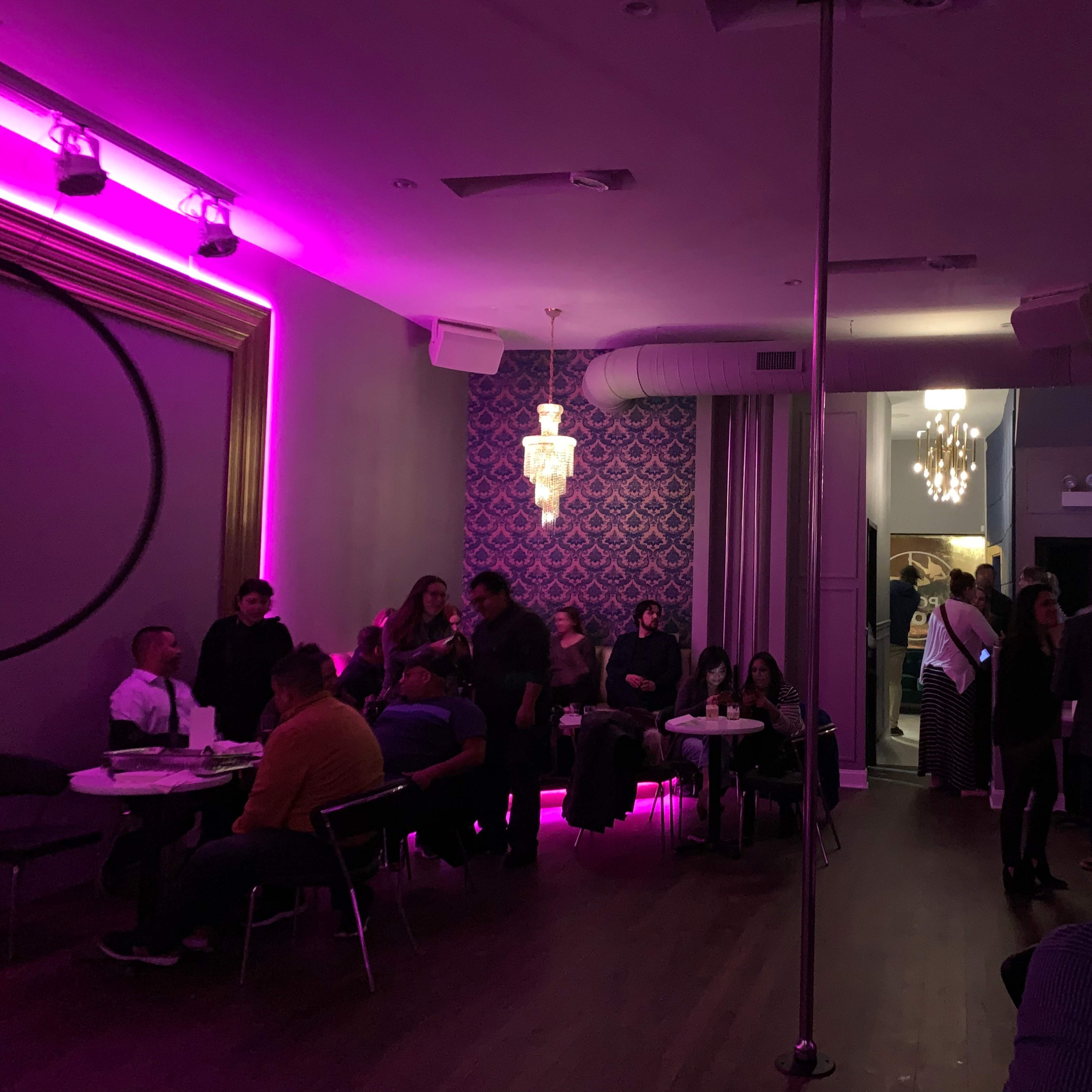 A lively indoor venue with pink lighting, featuring clusters of people socializing at tables and a pole in the center of the room.