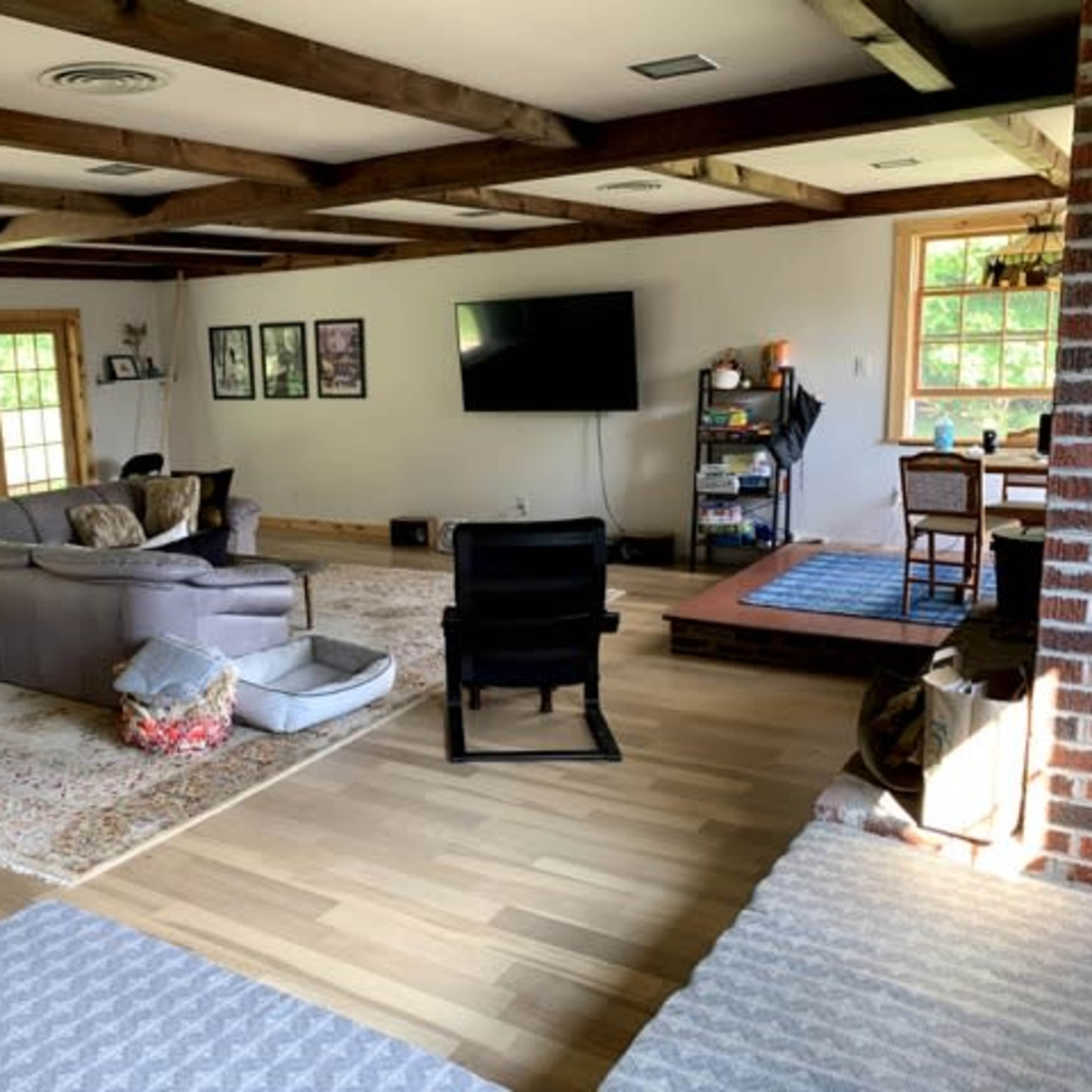 A spacious living room with wooden beams, a large sectional sofa, a television mounted on the wall, and a laid-back seating area near a window.