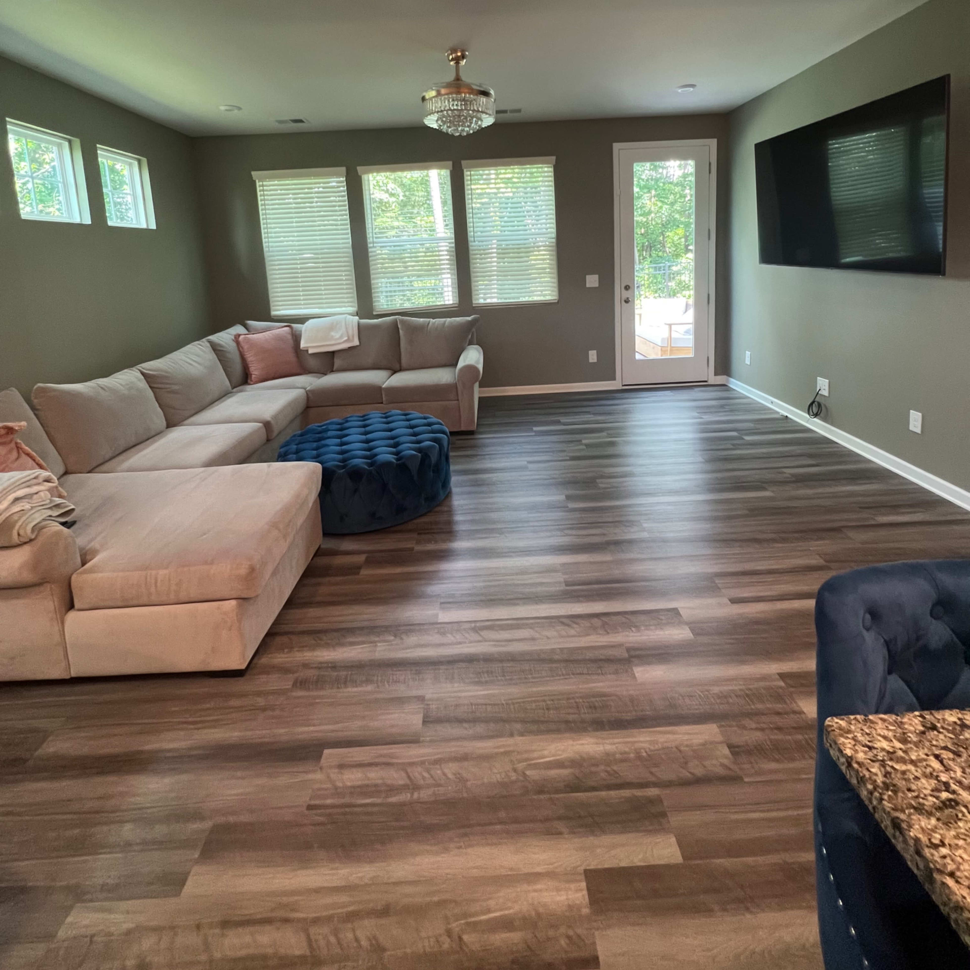 The living room features a sectional sofa, a round ottoman, a large TV, and windows that provide natural light.