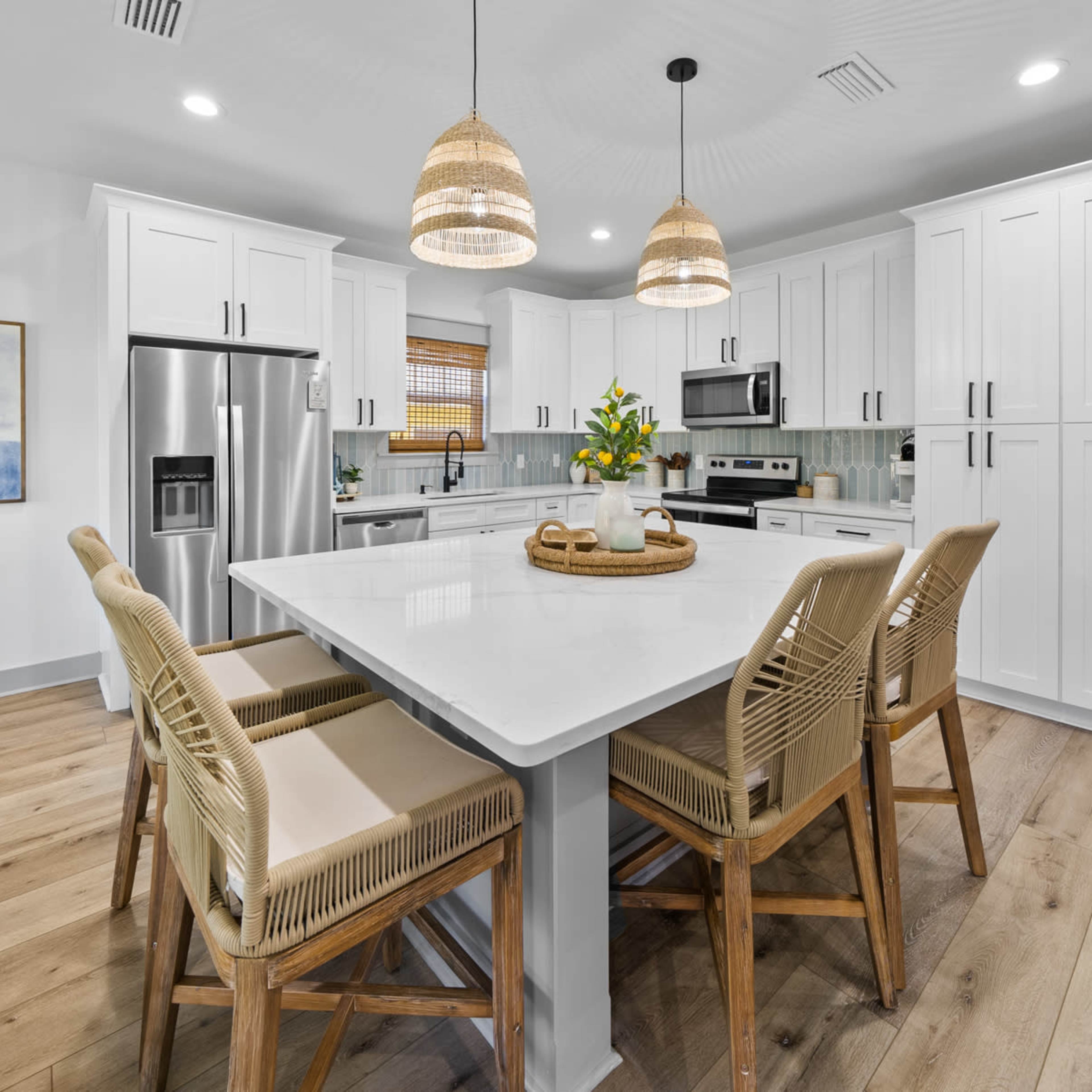 A modern kitchen features white cabinetry, stainless steel appliances, and a large island surrounded by four wicker-barred chairs.