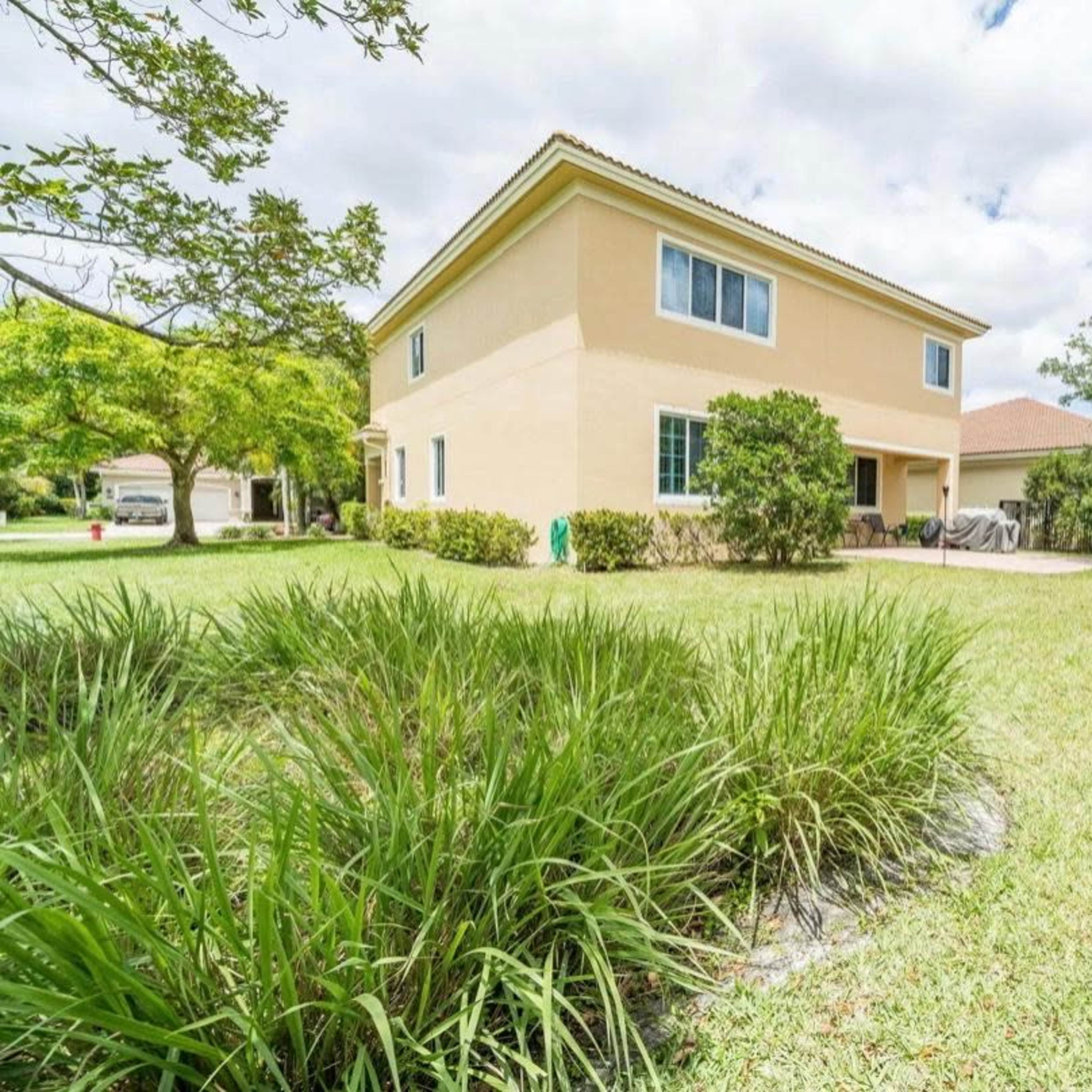 A two-story house with a patio is set in a grassy yard surrounded by bushes and trees.