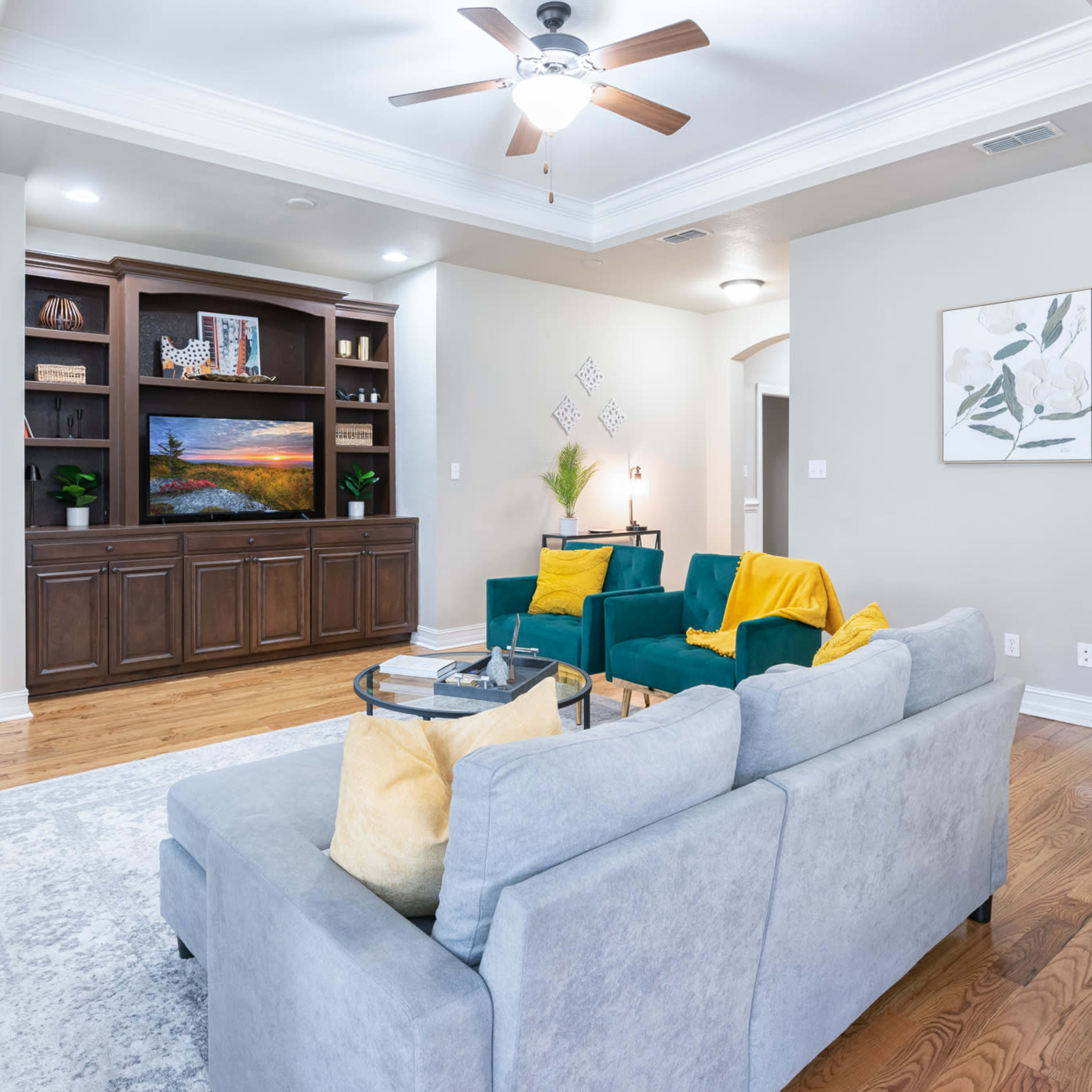 The living room features a light gray sofa, two teal chairs with yellow throws, a wooden entertainment center, and a large potted plant.