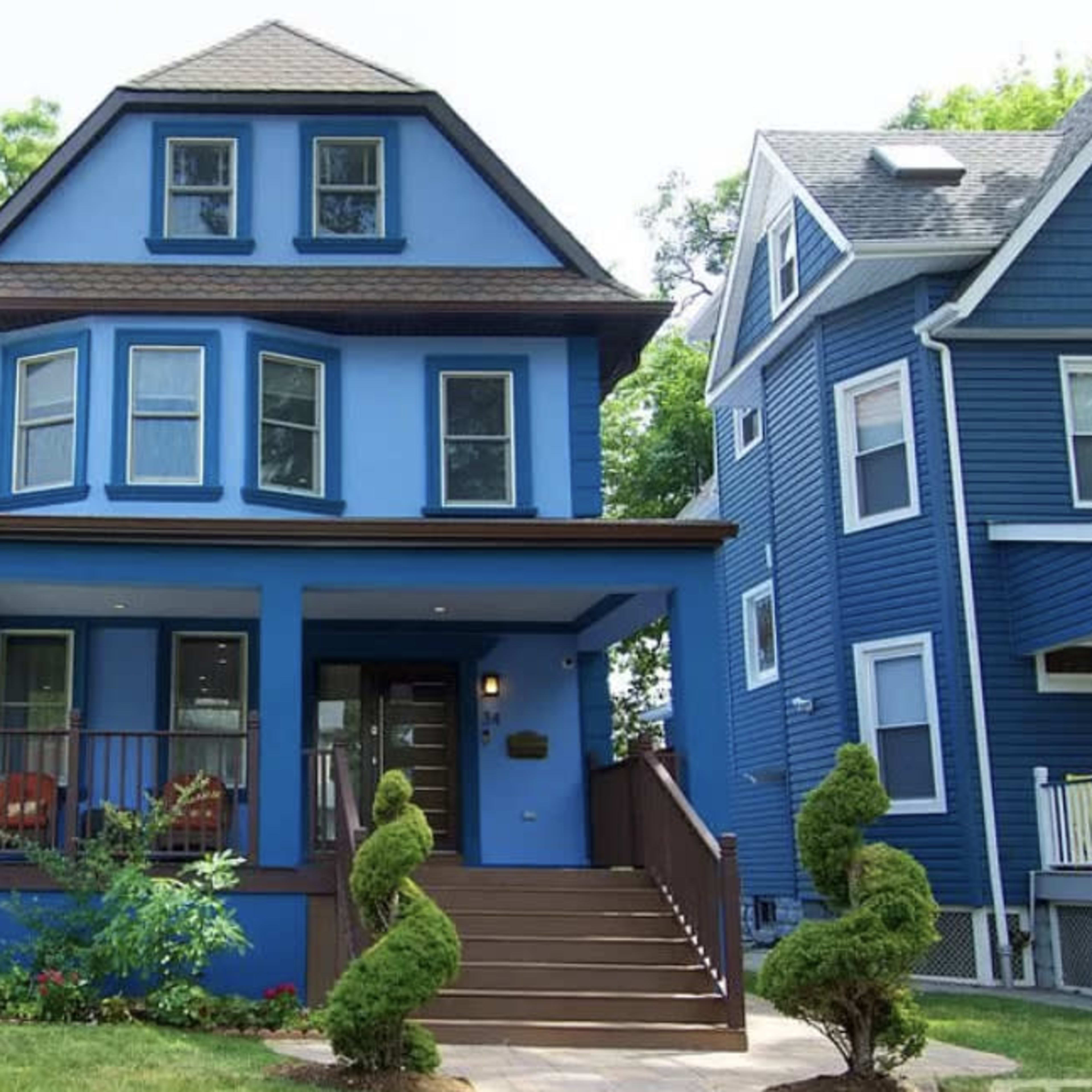 A blue three-story house with a front porch and landscaped steps is flanked by two other residential buildings.