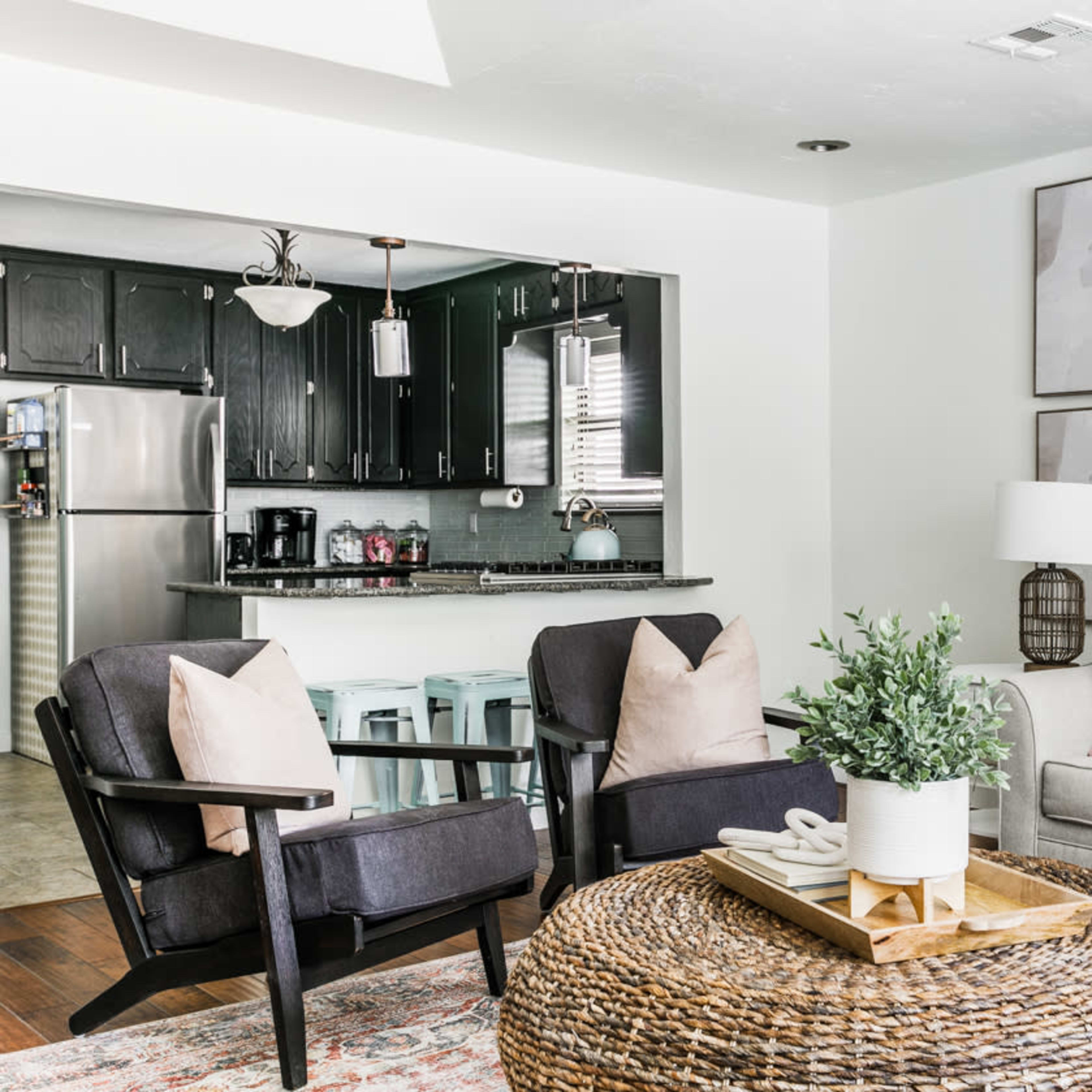 A modern living room with a gray sofa, two black armchairs, a round woven coffee table, and a kitchen area visible in the background.